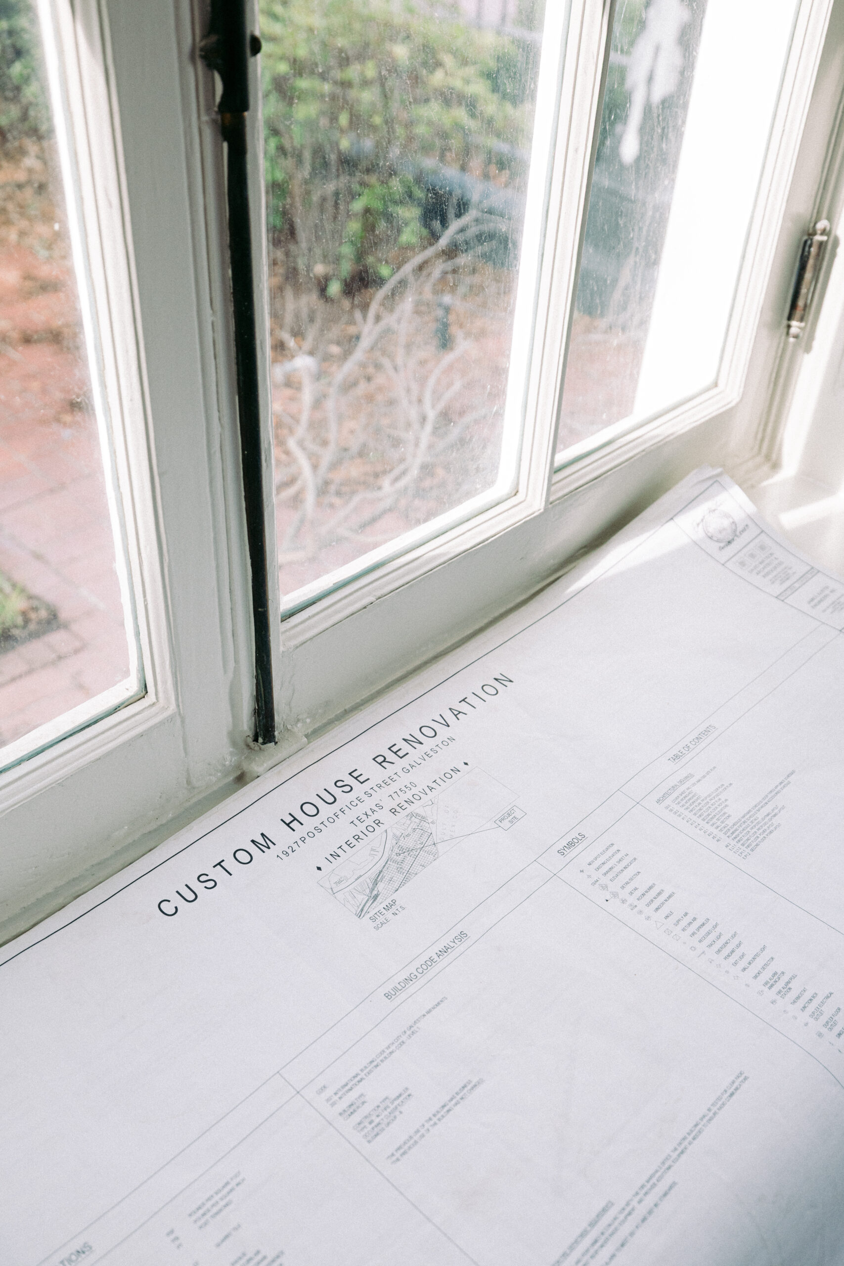 A set of custom house renovation blueprints is spread out on a windowsill beneath a window, with a garden visible outside. - McNatt Contracting