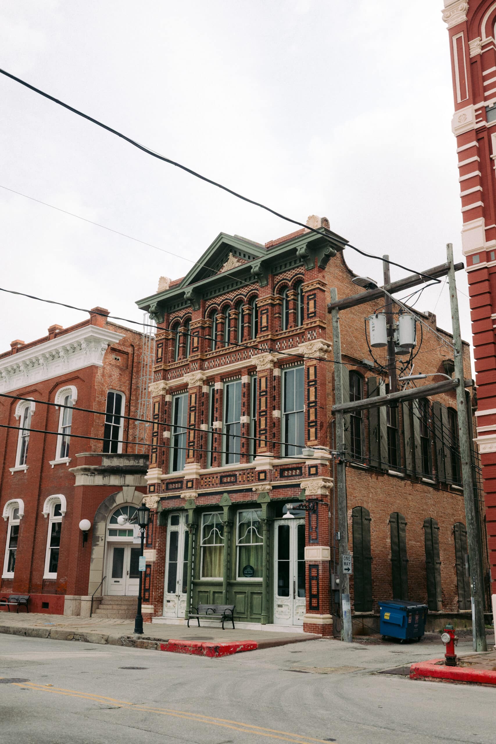 A two-story historic brick building with ornate architectural details, tall windows, and green trim stands on a street corner. Overhead utility wires cross in front. Other brick buildings are adjacent. - McNatt Contracting