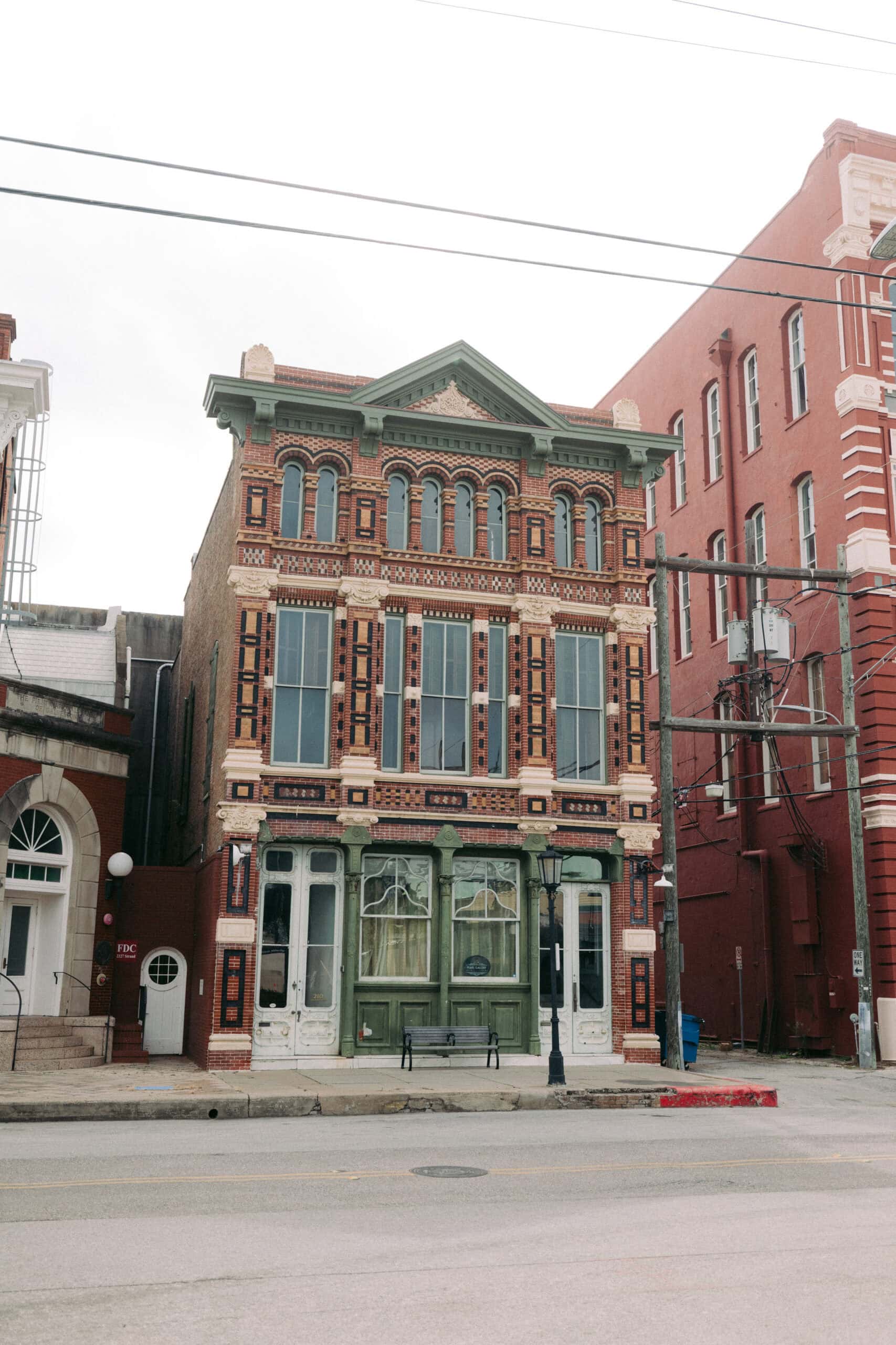 A three-story brick building with ornate architectural details and large front windows stands between taller red brick structures on a quiet city street. Overhead utility wires cross in front. - McNatt Contracting