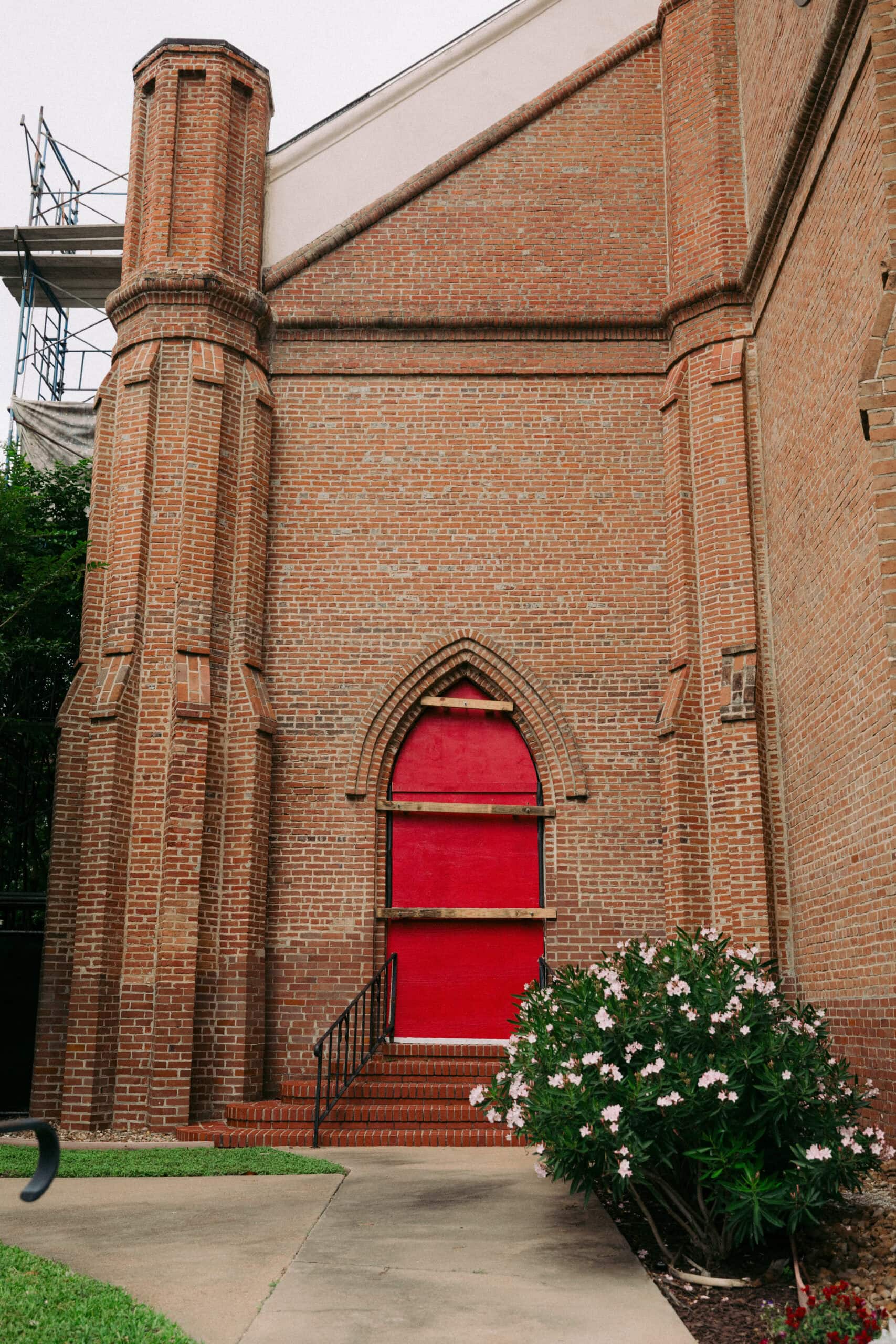 A red, arched door with steps leading up to it is set in a tall brick building. A small green bush with white flowers is in the foreground, and a concrete path curves along the lower edge of the image. - McNatt Contracting
