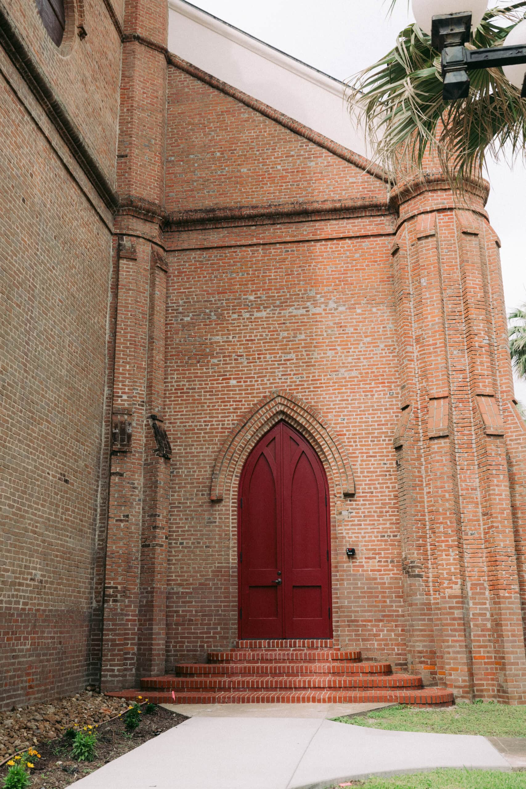 A tall, red, arched wooden door set in the brick exterior of a historic building. The doorway is raised with several steps leading up to it. There is a small patch of grass and flowers in the foreground. - McNatt Contracting