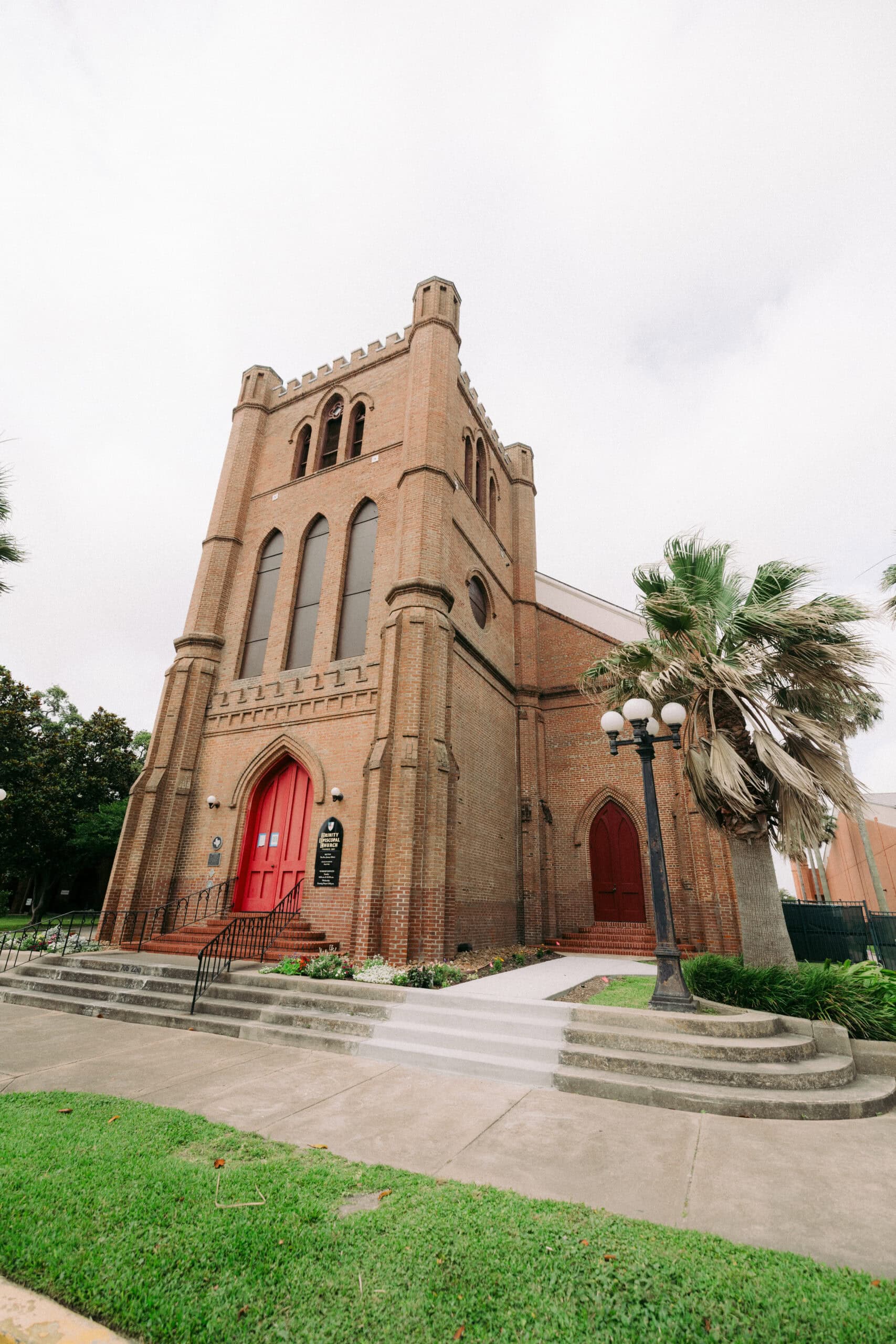 A large brick church with a tall tower, arched windows, and two red doors welcomes you. Stairs and a ramp lead to Trinity’s entrance. A lamppost and palm tree stand on the right, with green grass beneath a cloudy sky. - McNatt Contracting