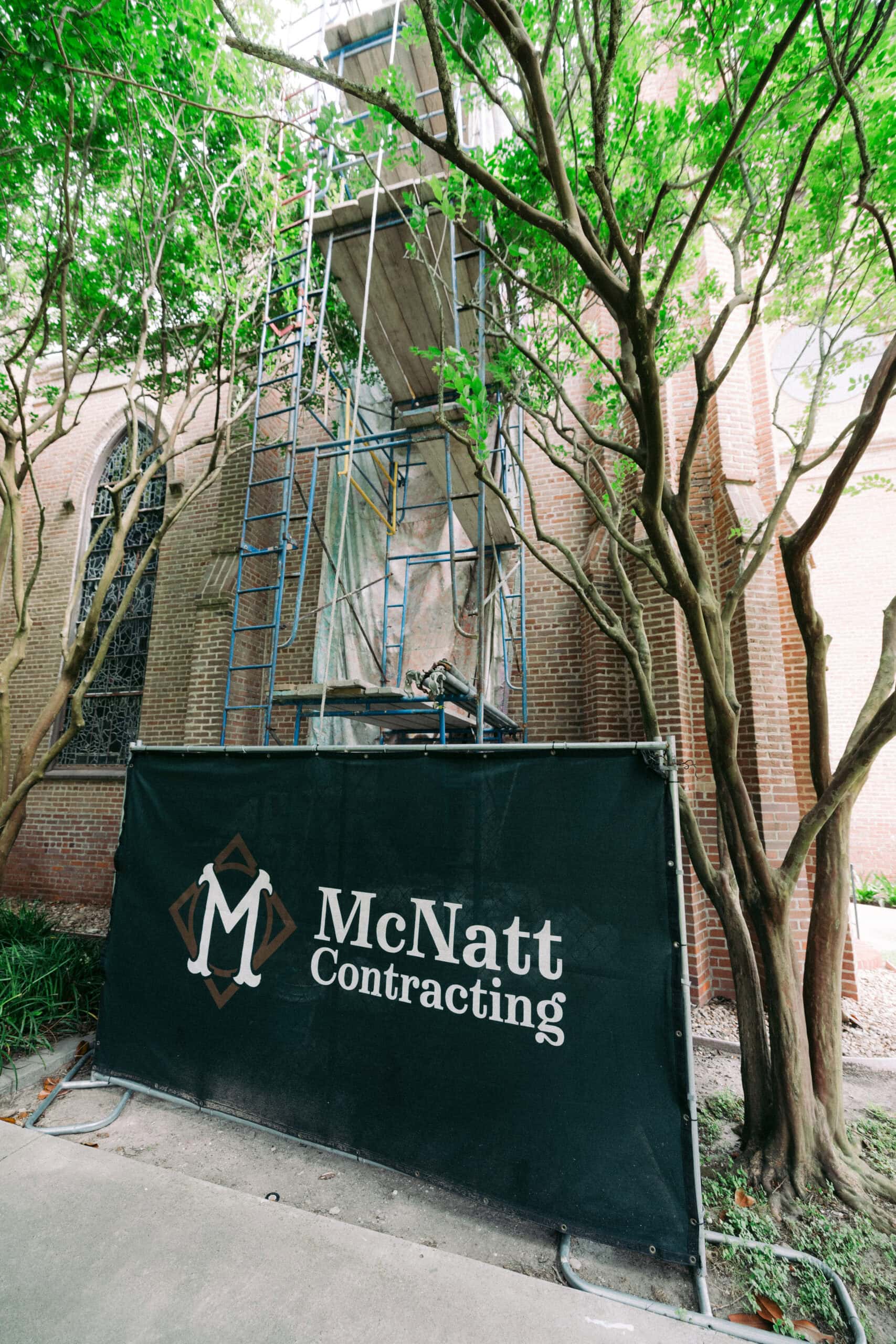 A construction site outside a brick building with scaffolding and a green barrier displaying the McNatt Contracting logo, near Trinity. Lush trees with green leaves surround the area. - McNatt Contracting