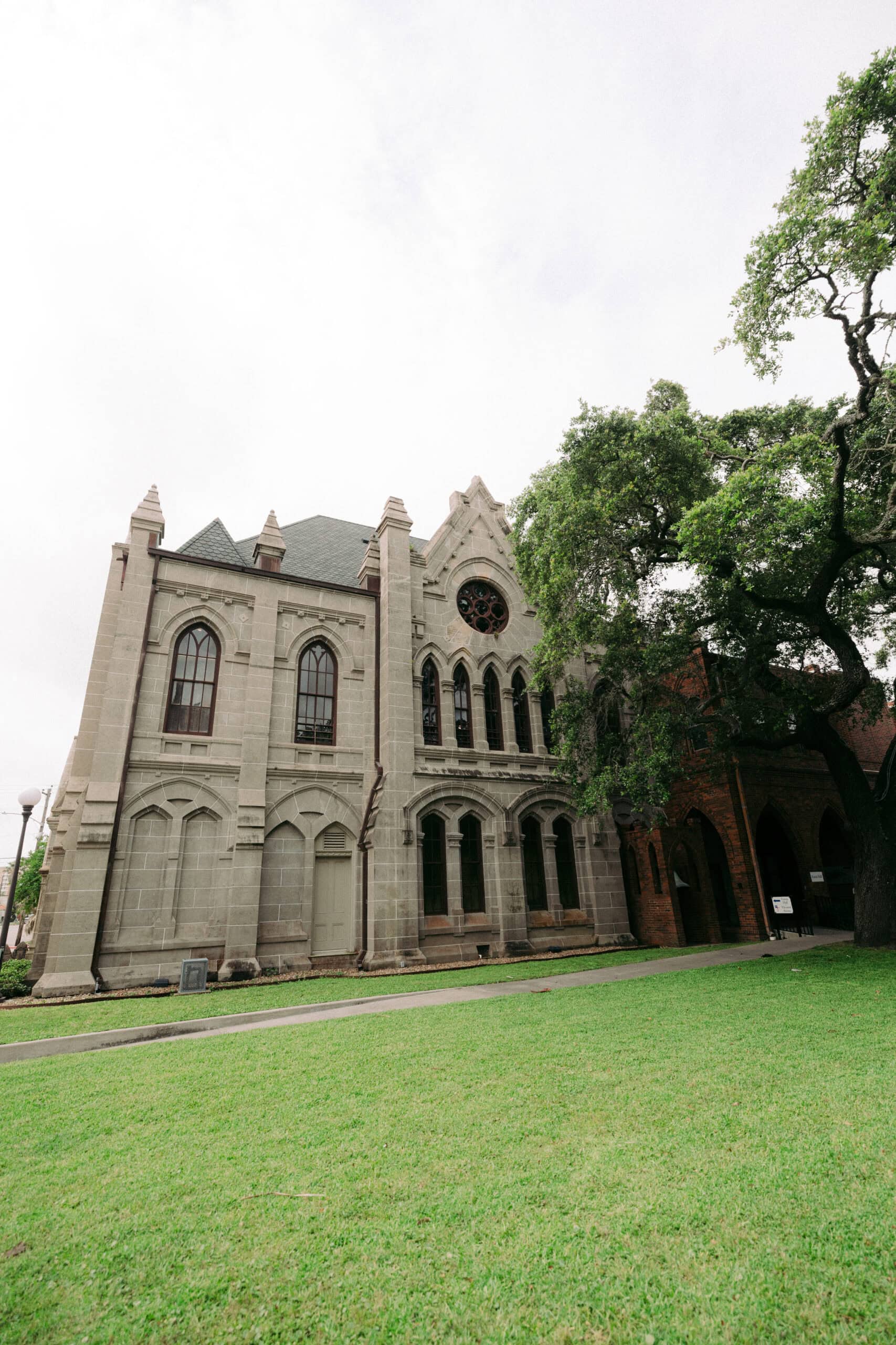 A stone building with arched windows and a rose window sits next to a large tree and a grassy lawn under an overcast sky. - McNatt Contracting