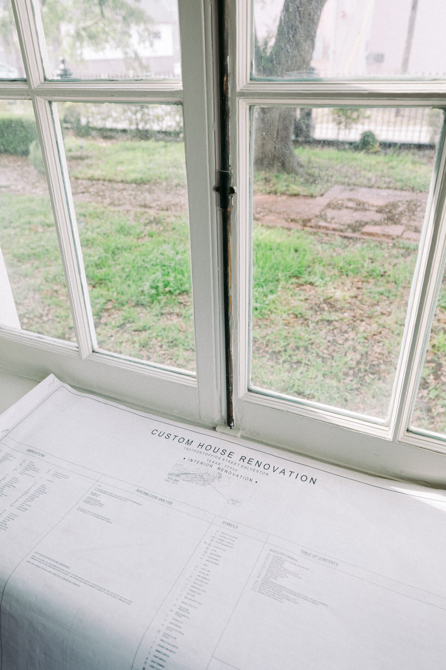 A set of architectural plans labeled "Custom House Renovation" rests on a windowsill inside a house, with a view of grass, trees, and a brick walkway outside the window. - McNatt Contracting