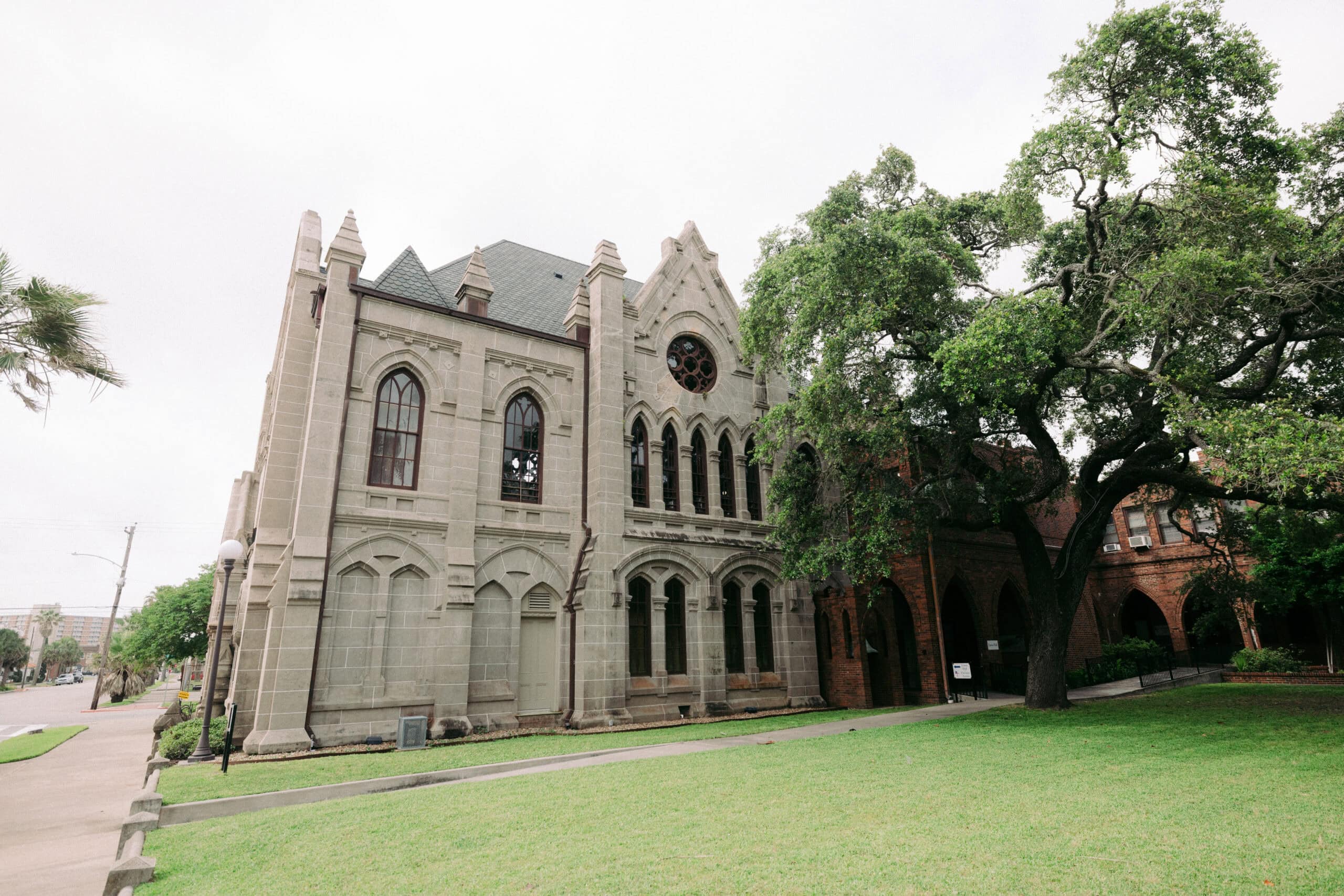 A historic stone building with pointed arches and a large circular window stands next to a lawn and large tree under a cloudy sky. The architecture features Gothic-style elements. - McNatt Contracting
