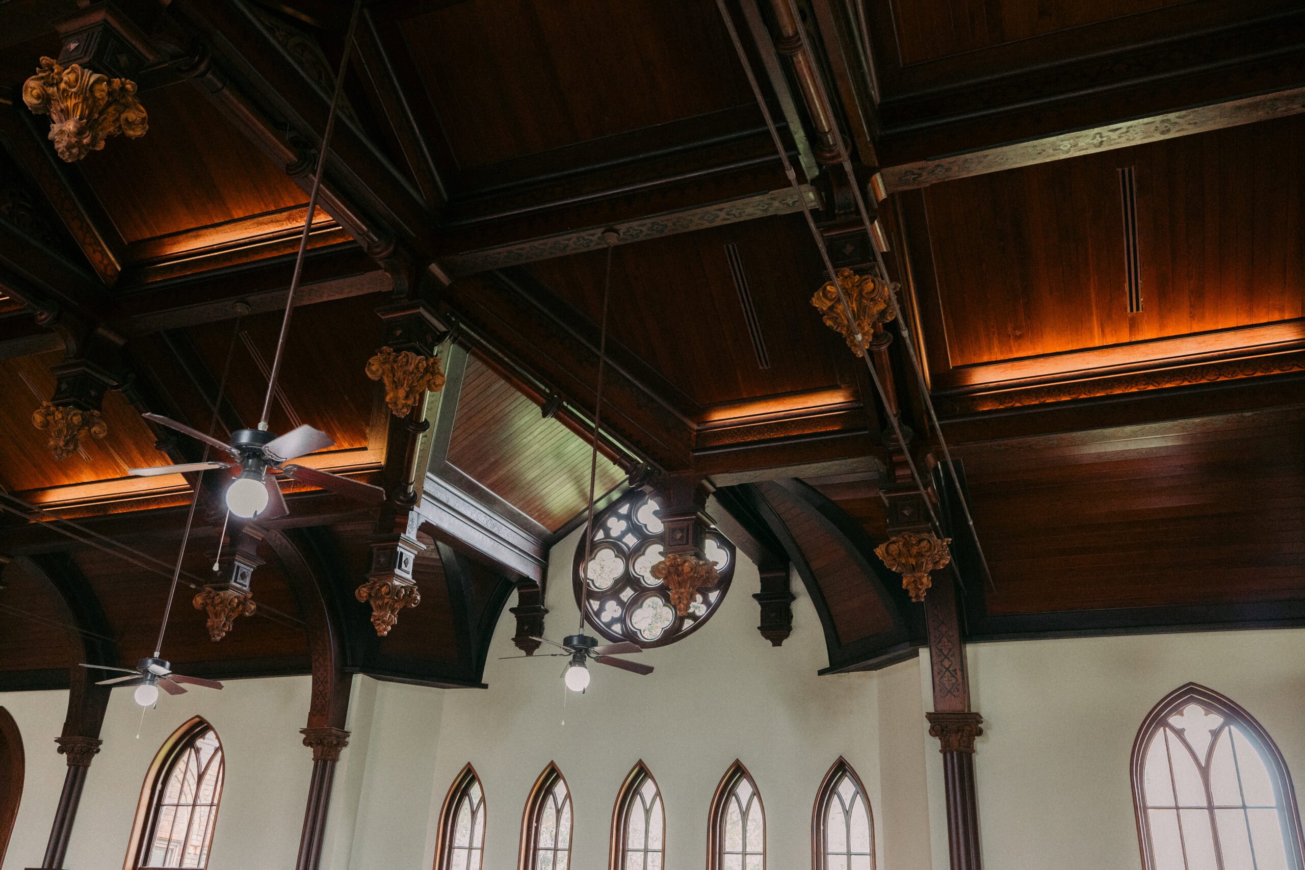 Wooden ceiling of a church interior featuring dark beams, decorative gold elements, ceiling fans, and stained glass windows along the white walls. - McNatt Contracting
