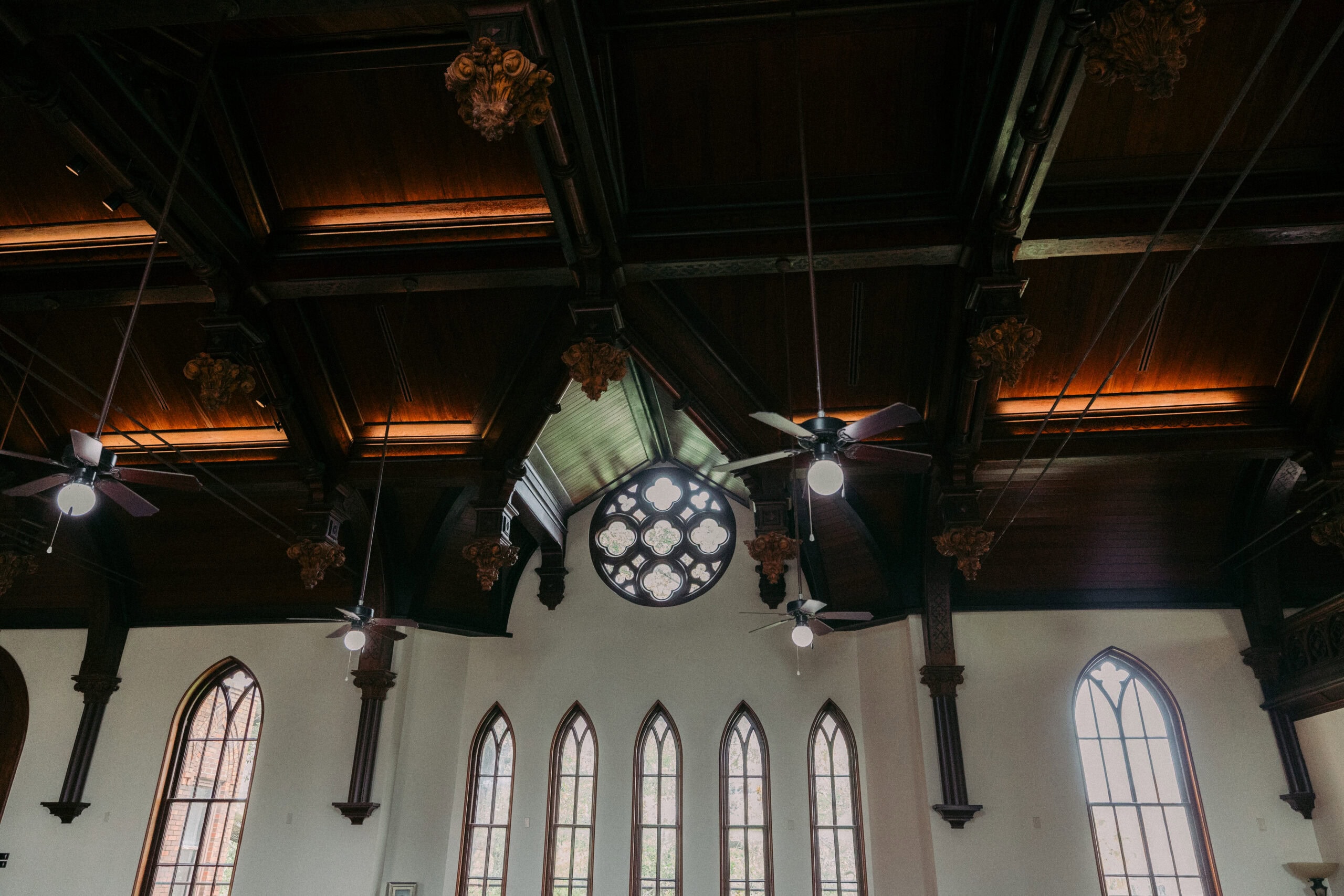 A view of the upper interior of a church, showing dark wooden vaulted ceilings, hanging ceiling fans, decorative beams, and tall arched windows with a circular stained glass window in the center. - McNatt Contracting