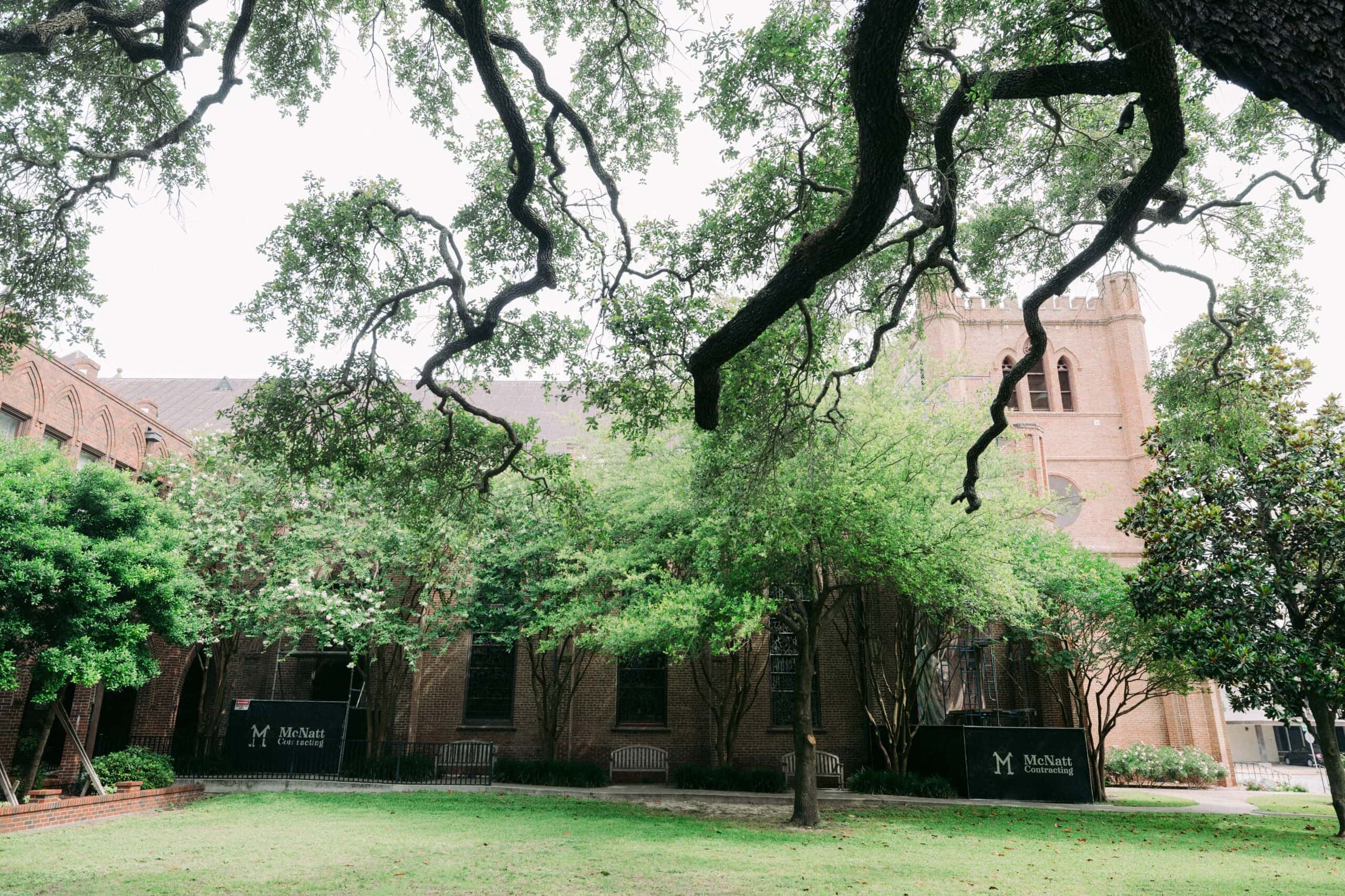 Large trees with sprawling branches frame a grassy courtyard in front of a red brick building. Black signs reading "McNay Art Museum" are visible near the building's arched windows and tower. - McNatt Contracting