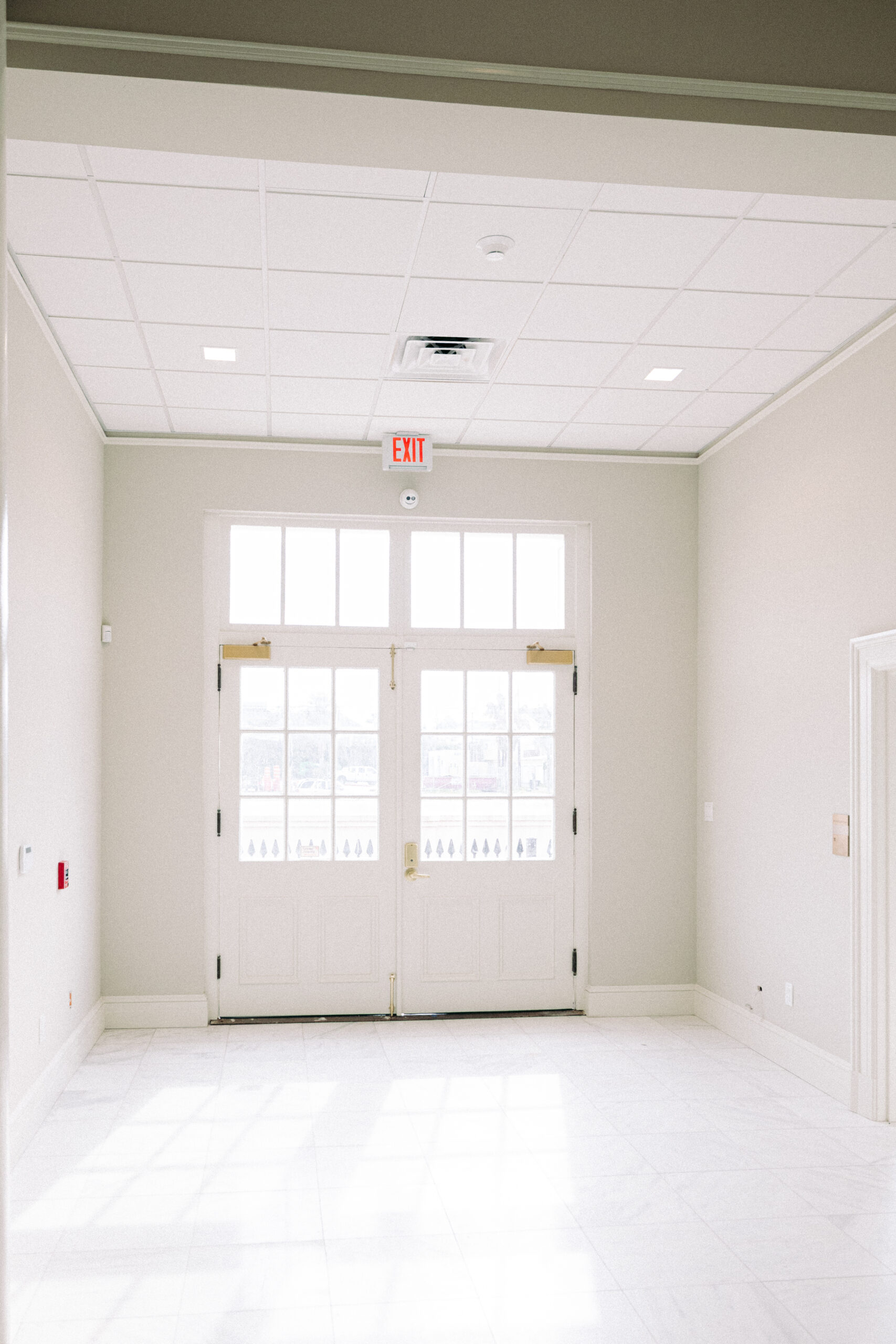 A bright, empty hallway with white walls and floor tiles, featuring double glass doors with sidelights at the end. An exit sign is above the doors, and a fire alarm is mounted on the left wall. - McNatt Contracting