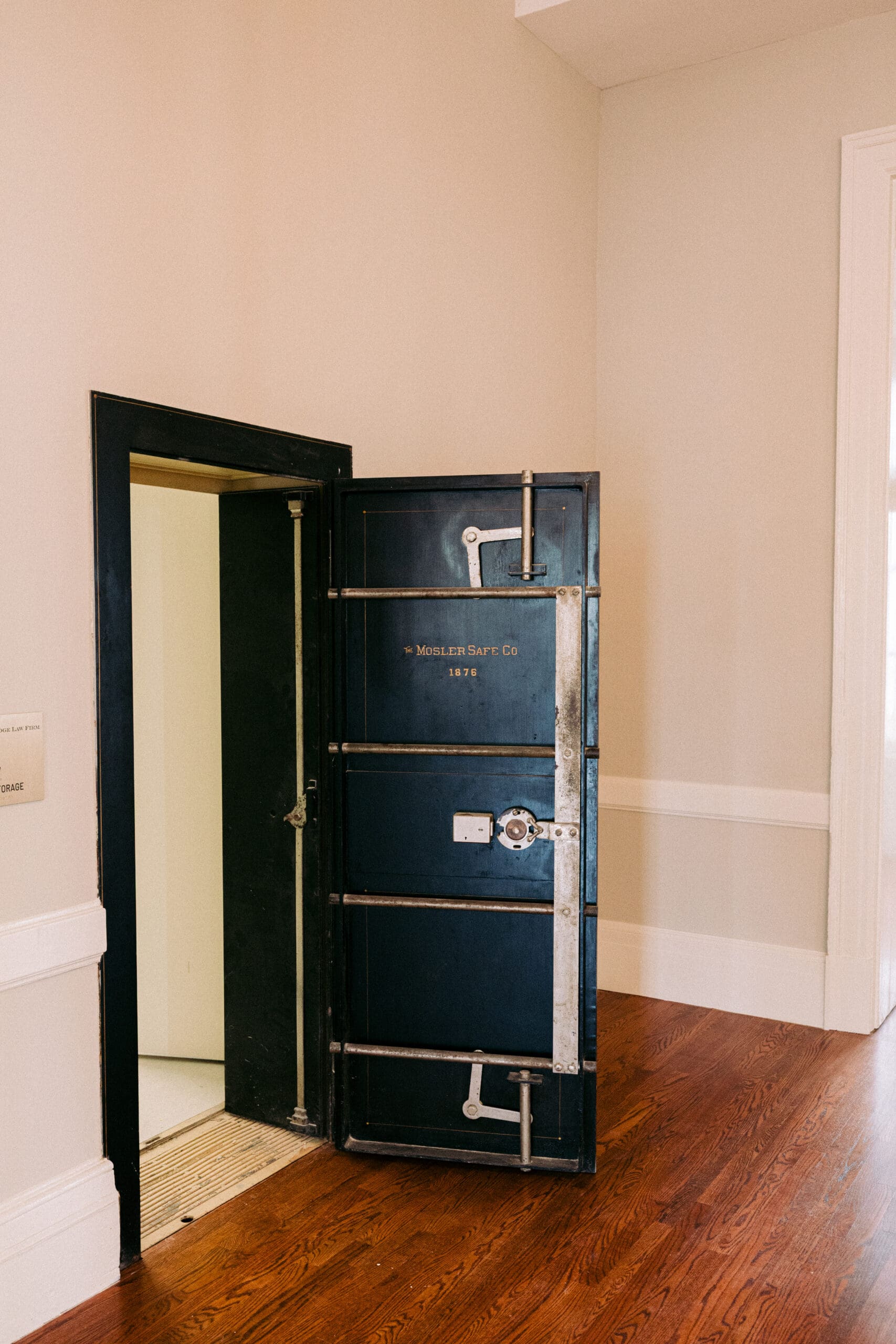 A large, open metal vault door labeled "Mosler Safe Co 1874" stands in a corner of a room with light-colored walls, white trim, and hardwood flooring. - McNatt Contracting