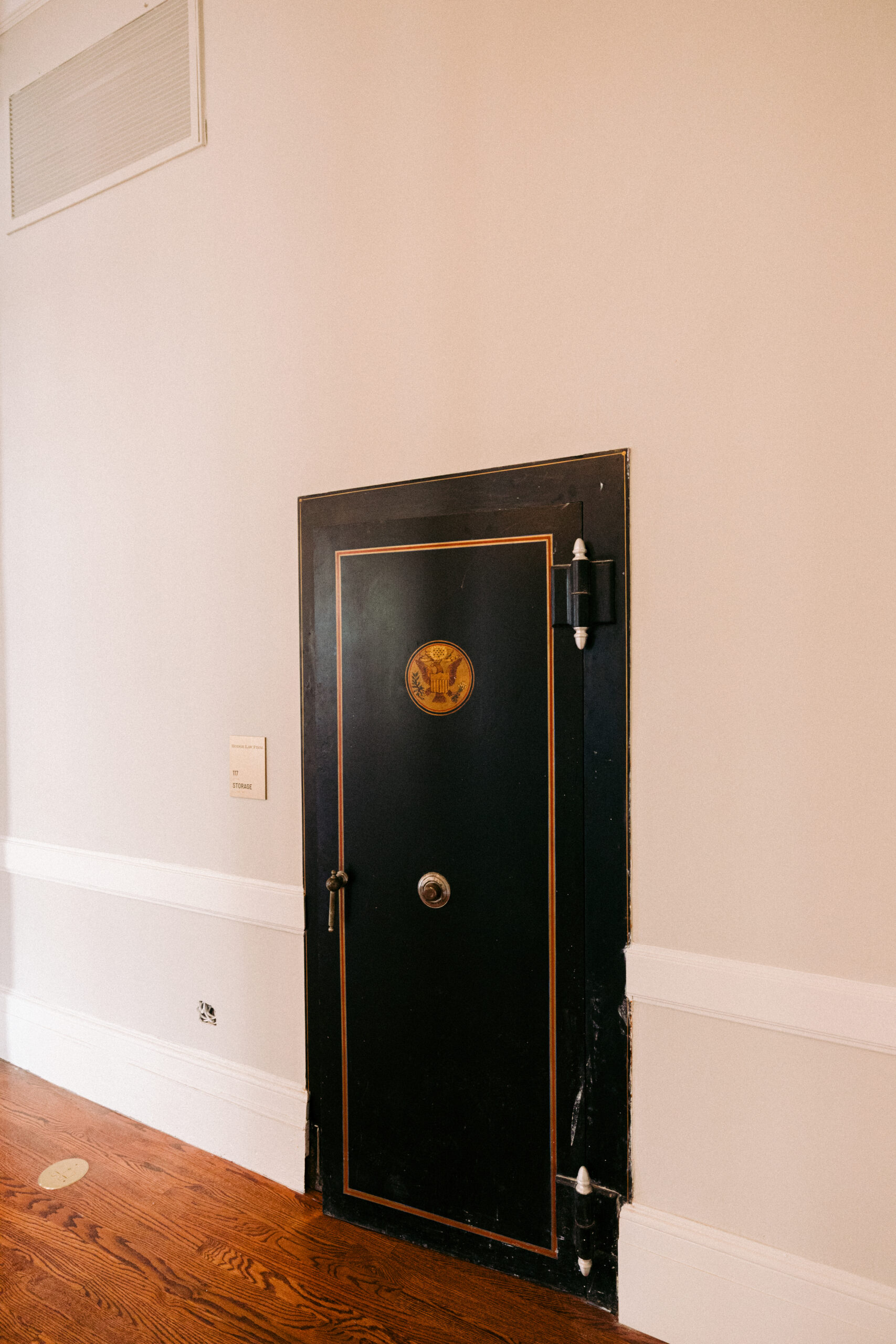 A large black metal door with a circular emblem and a round handle is set into a beige wall above a wooden floor. The door has gold trim and visible hinges on the right side. - McNatt Contracting