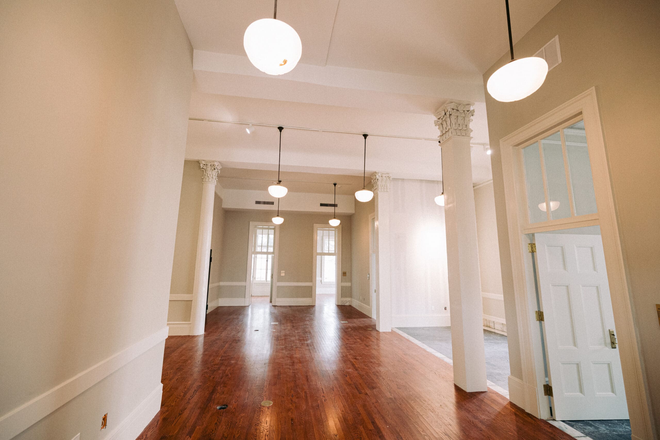 A spacious, empty room with high ceilings, white walls, hanging round light fixtures, decorative columns, wood flooring, and large windows letting in natural light. An open door is visible on the right side. - McNatt Contracting