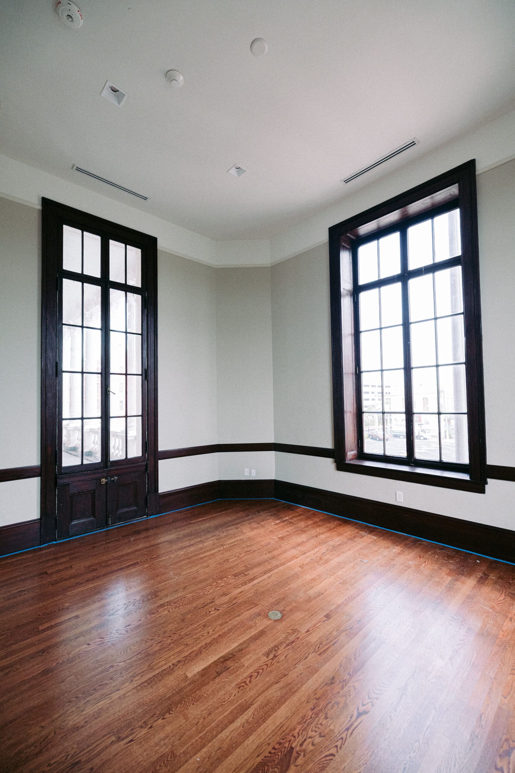 An empty room with hardwood floors, two large windows with dark wooden frames, beige walls, and white ceiling. There are electrical outlets on the walls and no furniture or decorations. - McNatt Contracting