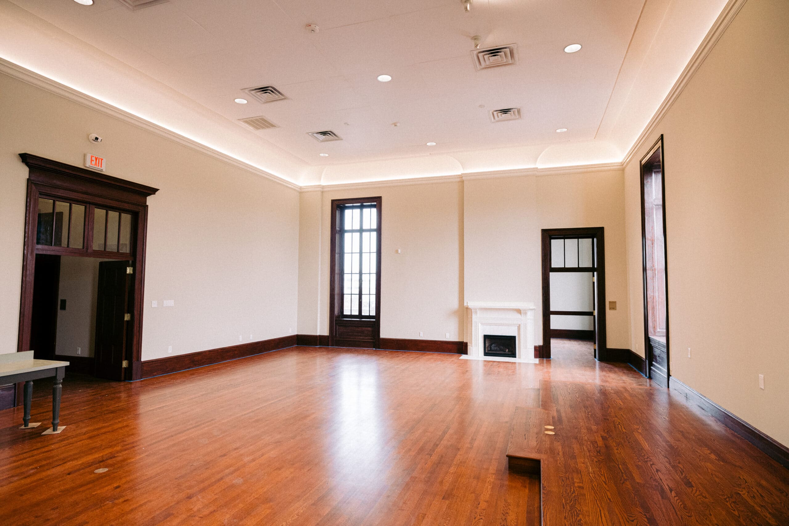 A spacious, empty room with polished wooden floors, high ceilings, a fireplace, tall windows, recessed lighting, and doorways leading to adjacent rooms. - McNatt Contracting