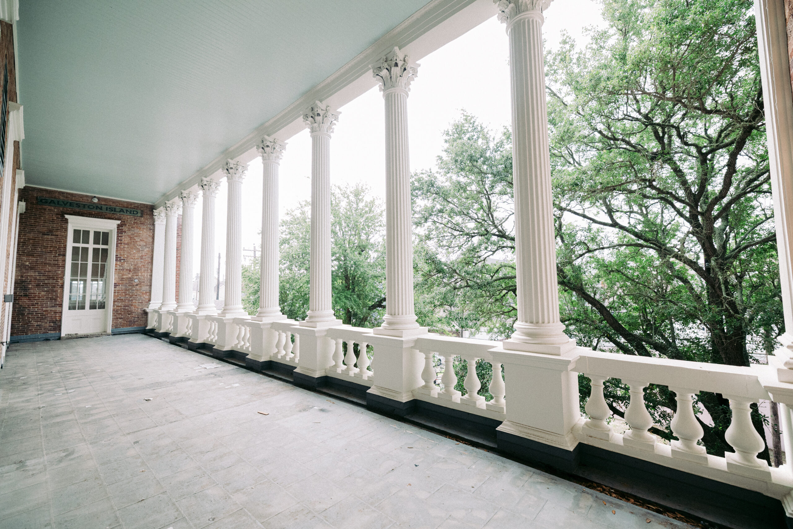 A long veranda with white columns and decorative railings, attached to a brick building, overlooks green trees on a cloudy day. The sign above the door reads "BRAVESTON HALL. - McNatt Contracting