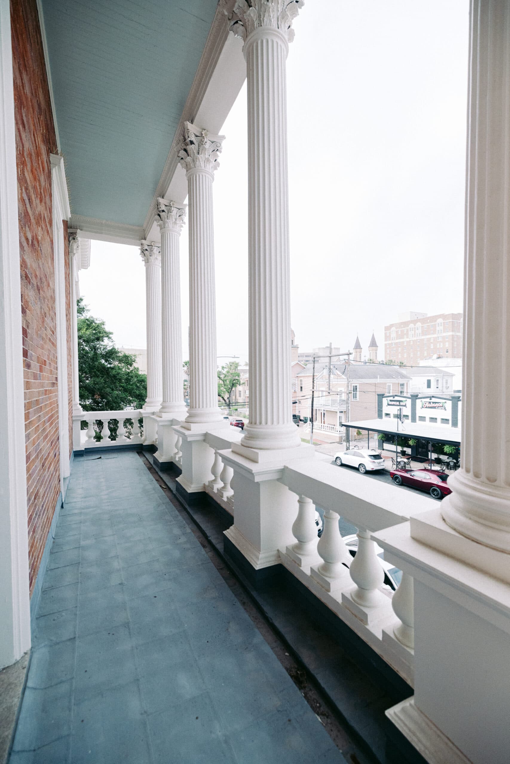 A long balcony with tall white columns, a pale blue ceiling, and a white balustrade overlooks a city street with buildings, parked cars, and trees visible in the background. - McNatt Contracting