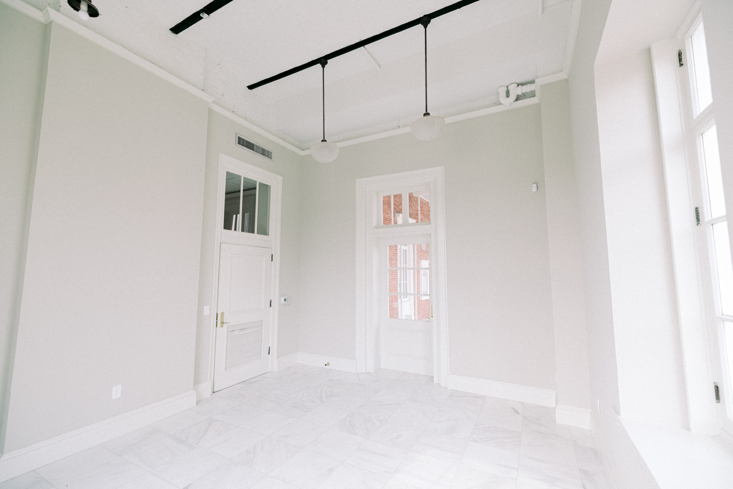 A bright, empty room with white walls, white tiled floor, large windows, a white door, and modern ceiling light fixtures. Natural light enters through the windows on the right. - McNatt Contracting