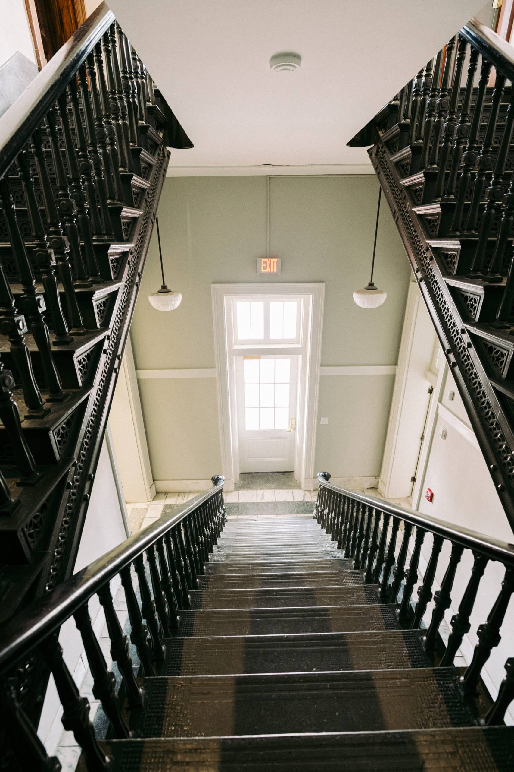 A view looking down a wooden staircase with ornate dark railings, leading to a white door with glass panels and an exit sign above it. The walls are light-colored and two round lights hang from the ceiling. - McNatt Contracting