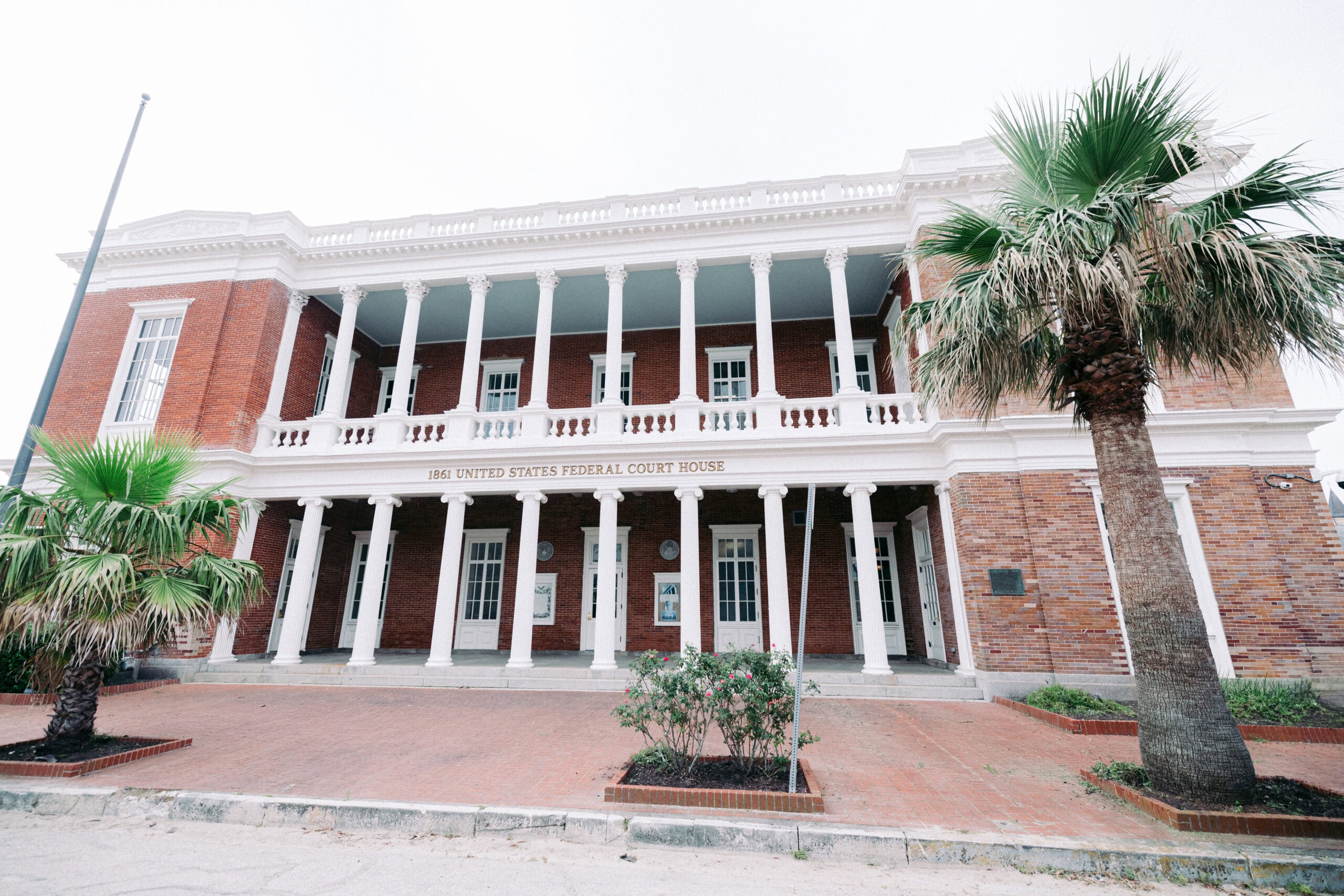 A two-story brick building with white columns at the entrance, labeled "United States Federal Court House." Palm trees and small shrubs are in front of the structure. - McNatt Contracting