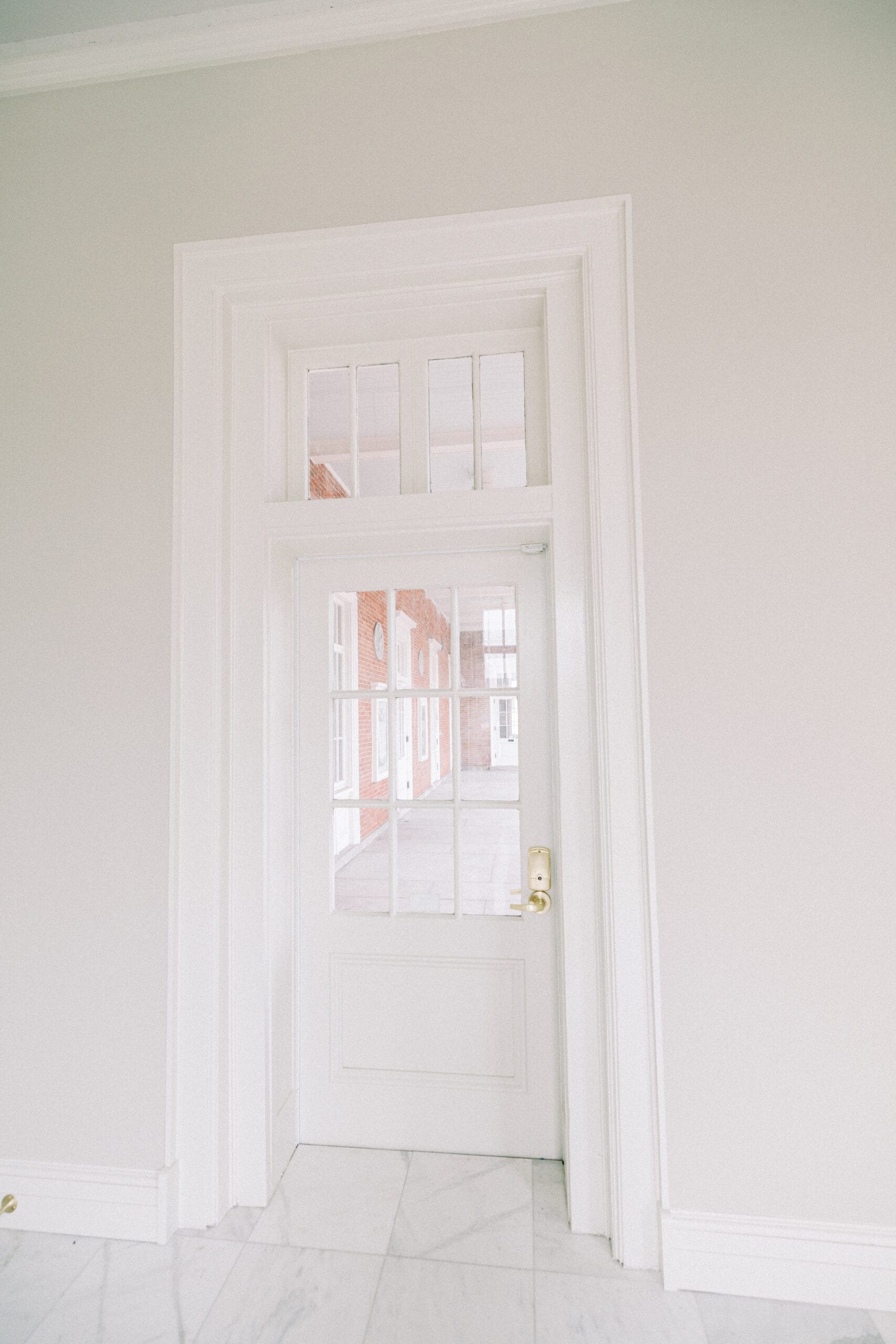 A white door with glass panels is set in a white wall, allowing a view of a hallway with white and red brick walls and large windows. The floor is white tile, and the door has a gold handle. - McNatt Contracting