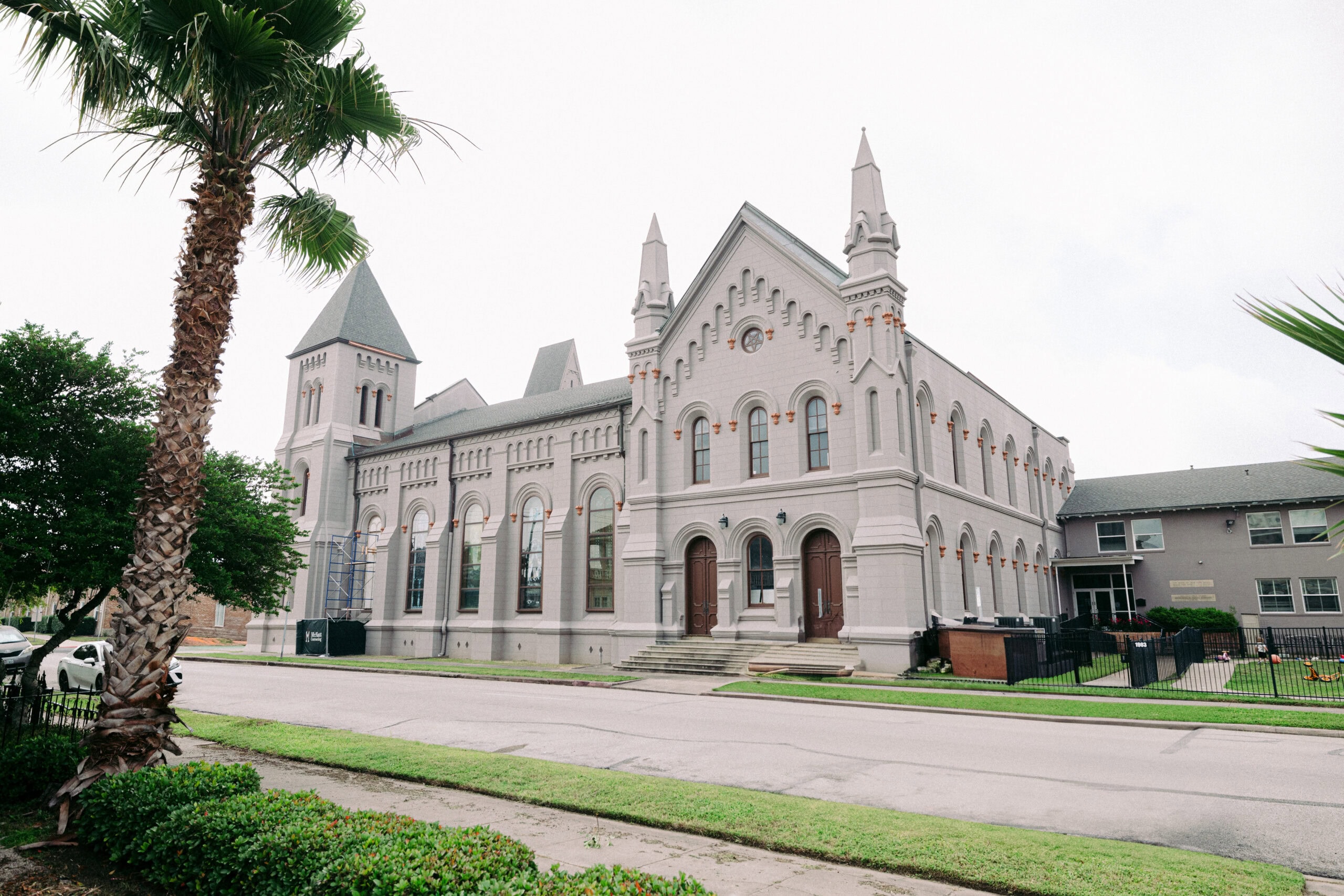 A large, light gray historic building—identified as First Presbyterian Church—with arched windows, pointed towers, and decorative trim sits near a palm tree along a suburban street on a cloudy day. - McNatt Contracting
