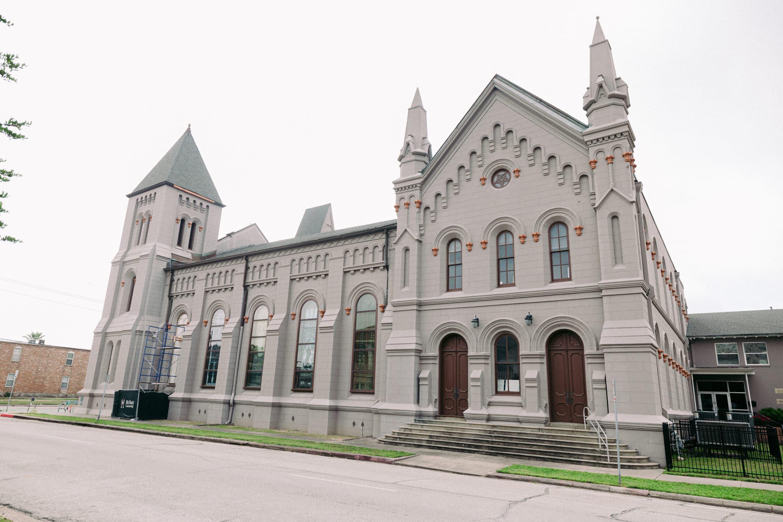 A large, historic-looking gray building with arched windows, twin towers, and decorative trim, seen from across a street with steps leading up to double wooden doors. A scaffold is visible on the left side of the building. - McNatt Contracting