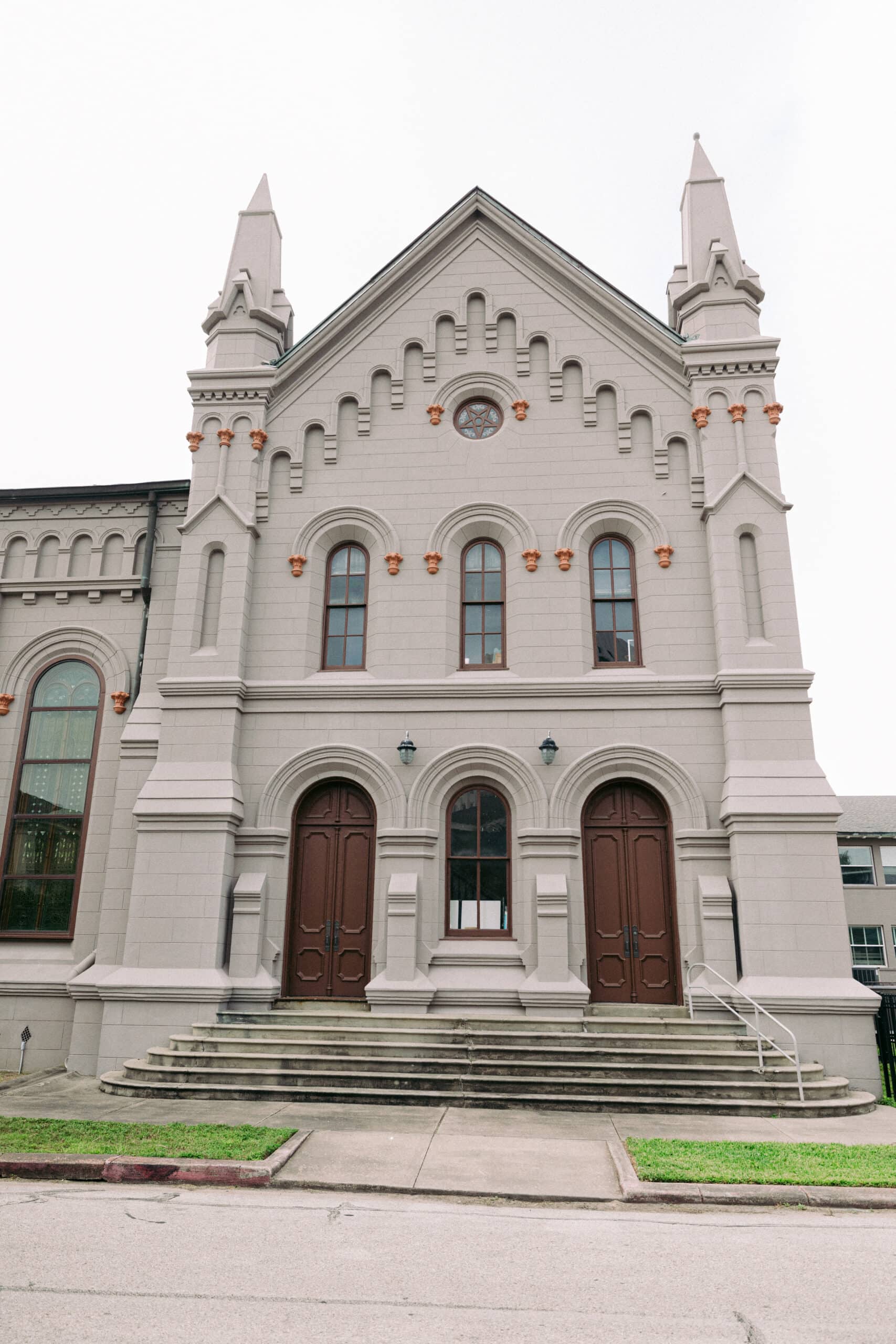 A light gray stone building with arched windows, three large wooden doors, and two tall pointed towers, viewed from the front. Steps lead up to the entrance. Grass and sidewalk are in the foreground. - McNatt Contracting