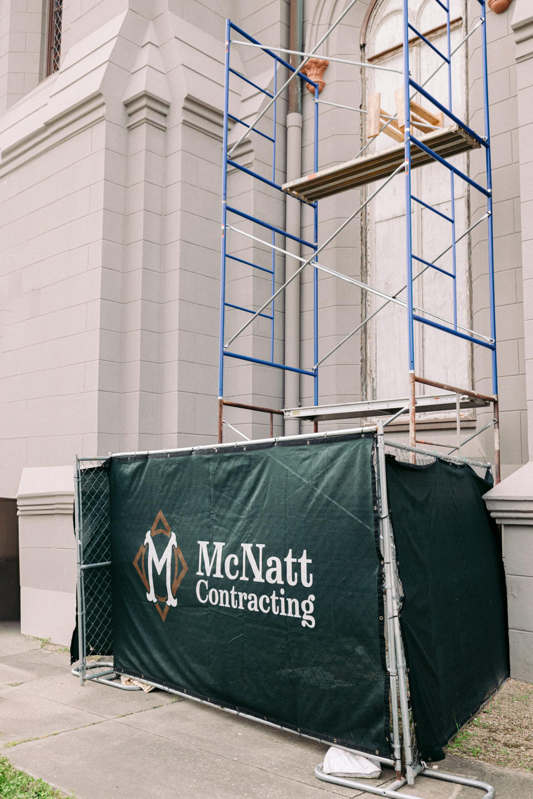 A construction scaffold stands against a building, enclosed by a green fence cover displaying the "McNatt Contracting" logo and name in white and gold lettering near the First Presbyterian Church. - McNatt Contracting