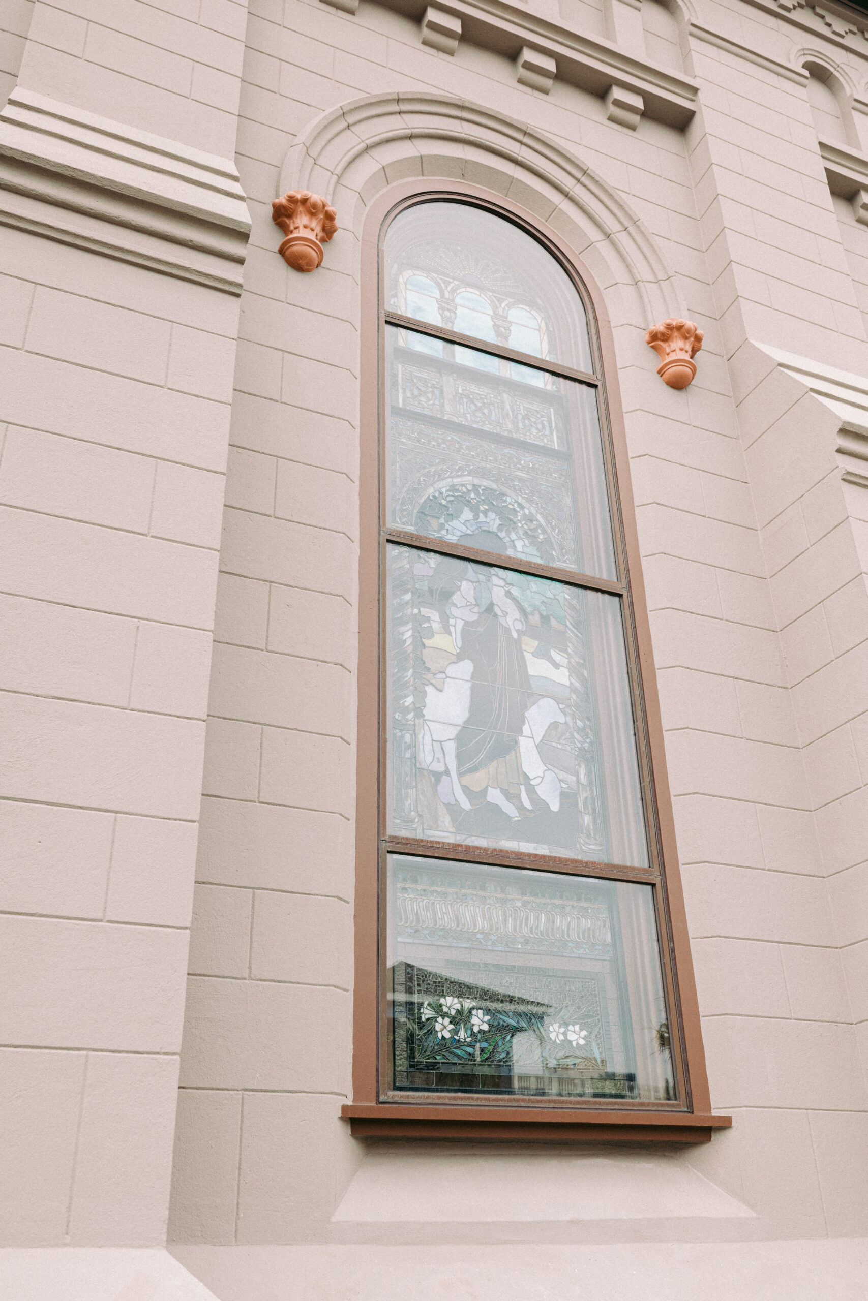 Tall arched window with stained glass depicting two figures, set in a beige stone building with decorative details and two small orange ornaments above the window. - McNatt Contracting