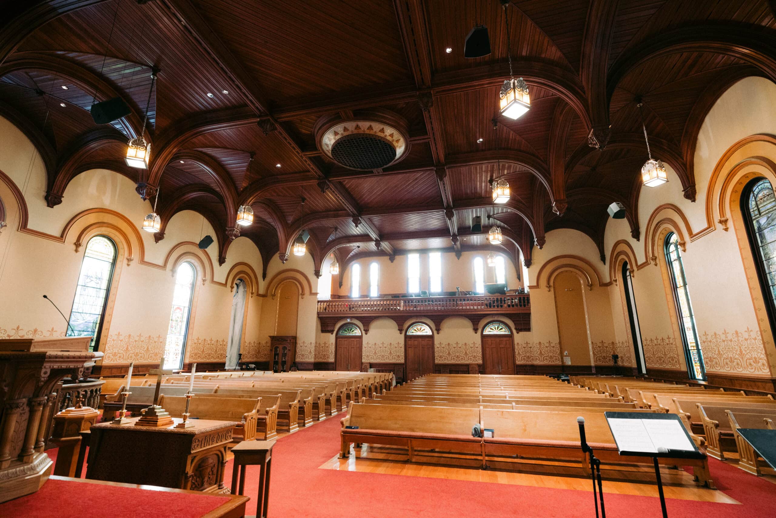 Interior of a large church with wooden pews, stained glass windows, ornate wooden ceiling, and a red carpeted aisle; the room is empty and well-lit with hanging lights. - McNatt Contracting