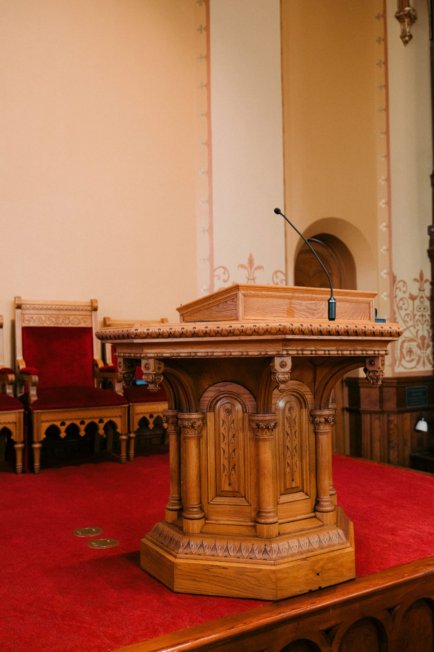 A wooden podium with a microphone stands on a red carpeted platform in First Presbyterian Church, surrounded by ornate woodwork and decorative walls. An empty, ornate wooden chair with a red cushion is in the background. - McNatt Contracting