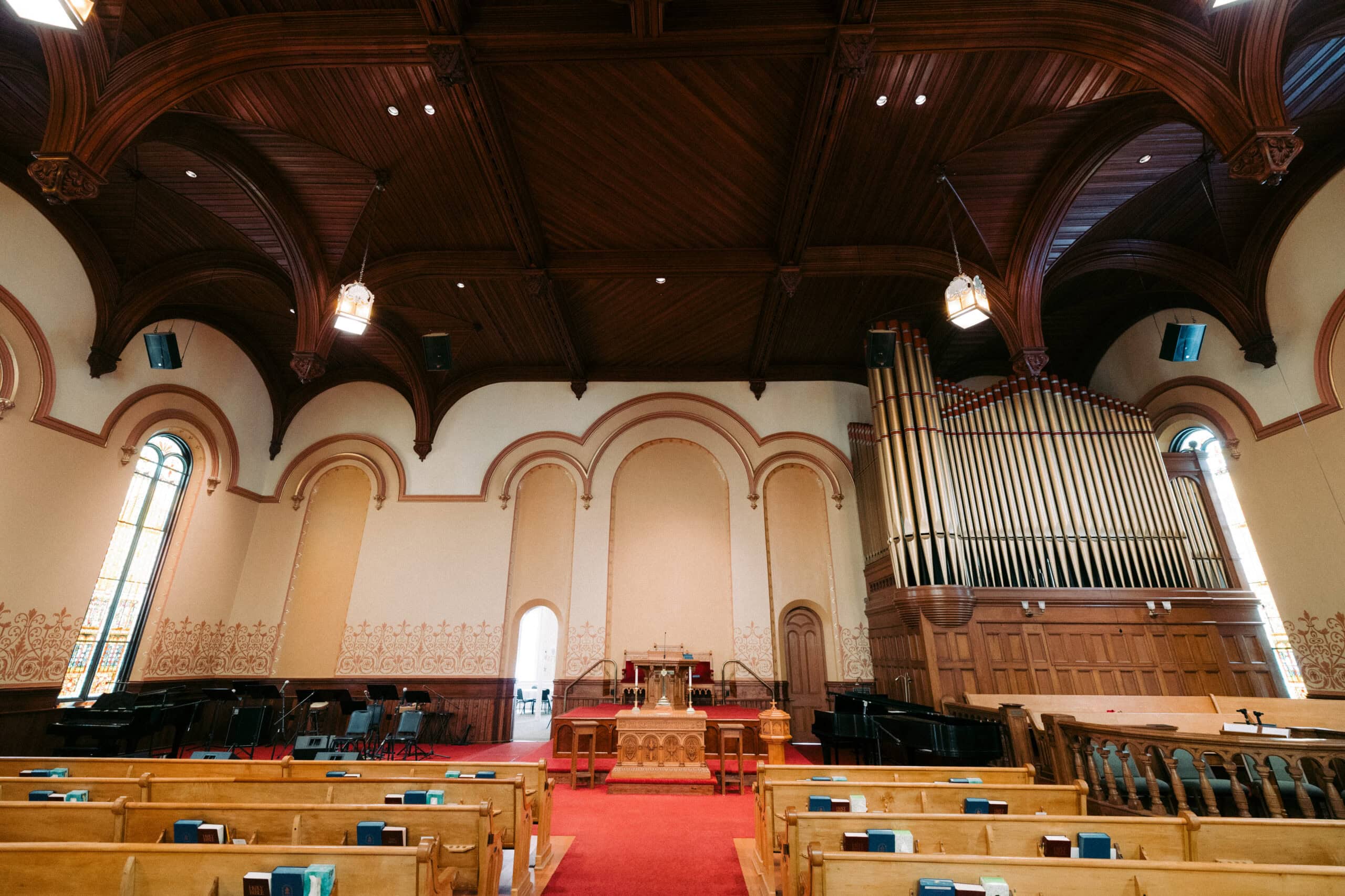 Interior of a church with wooden pews, a red carpeted center aisle, an altar at the front, tall pipe organ on the right, arched stained glass windows, and wooden vaulted ceiling with hanging lights. - McNatt Contracting