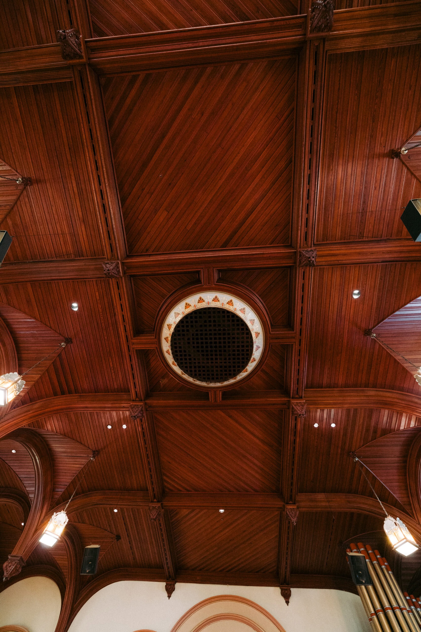 Wooden coffered ceiling with a large circular vent in the center, illuminated by hanging lamps and small recessed lights, and partial views of walls and organ pipes at the edges. - McNatt Contracting