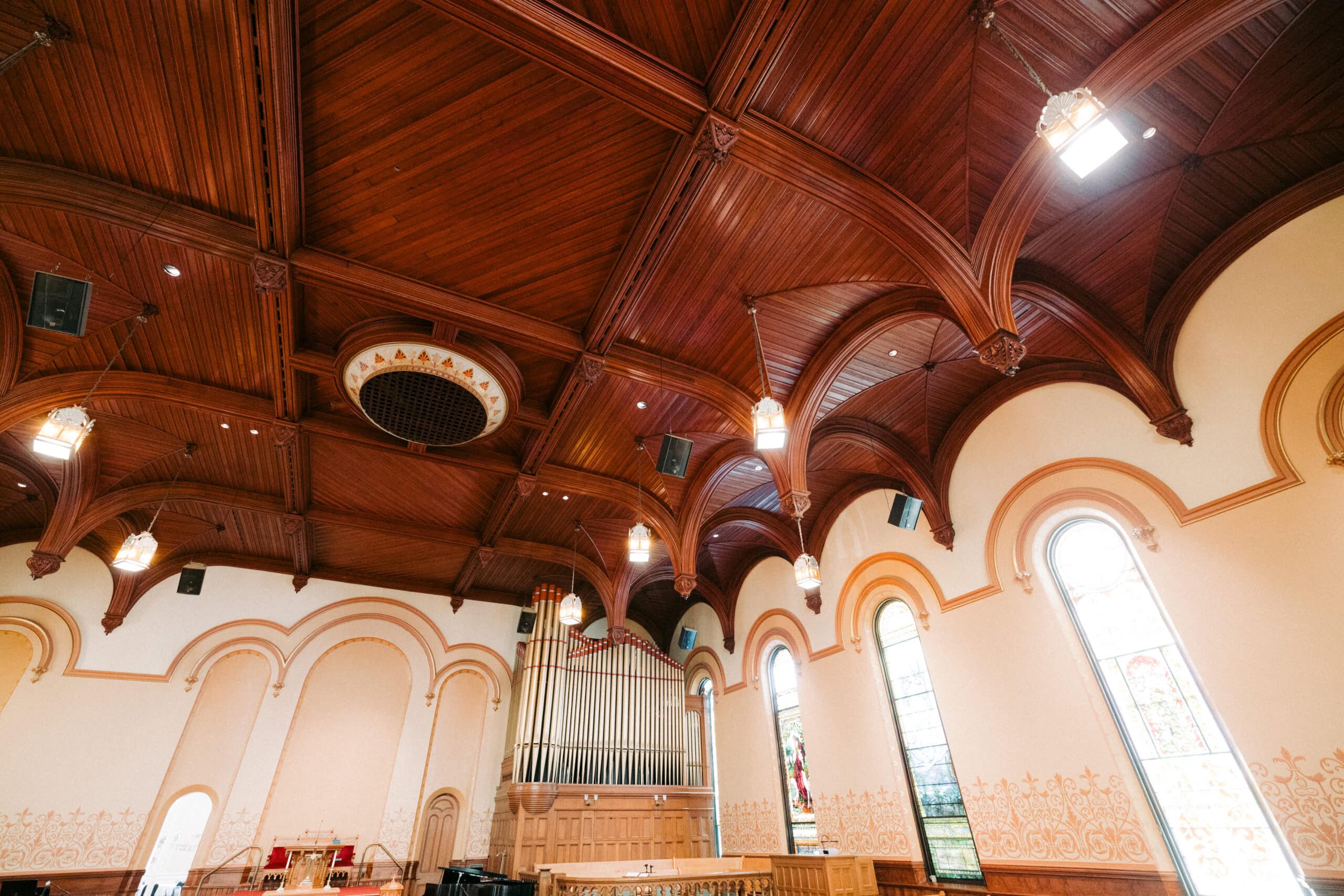 The image shows the interior of First Presbyterian Church with a high, ornate wooden ceiling, arched stained glass windows, a large pipe organ, and decorative wall detailing. Warm lighting hangs from the ceiling. - McNatt Contracting