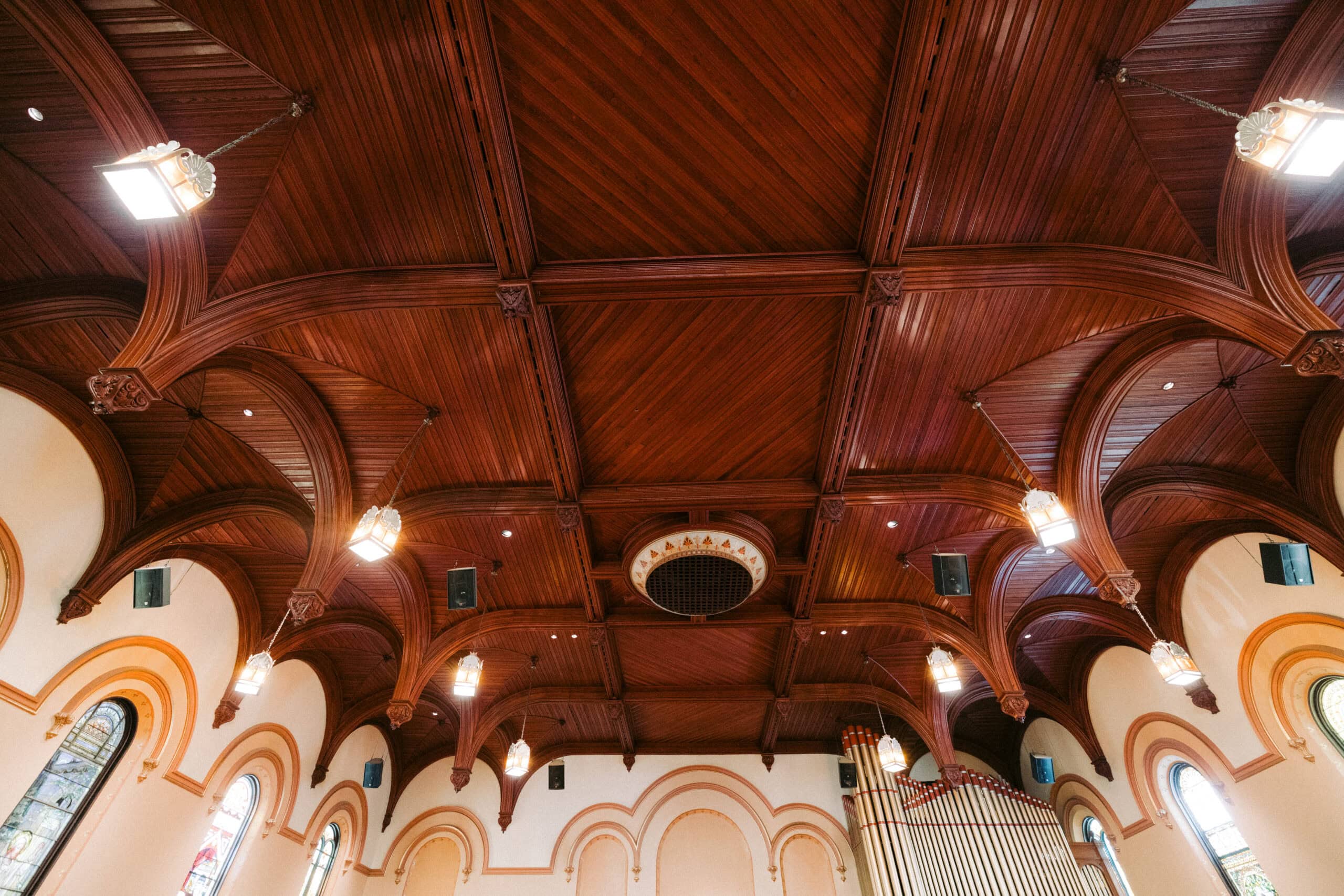 The interior ceiling of a church featuring wooden paneling, arches, hanging light fixtures, and stained glass windows along the walls. A circular vent or decorative element is in the center of the ceiling. - McNatt Contracting