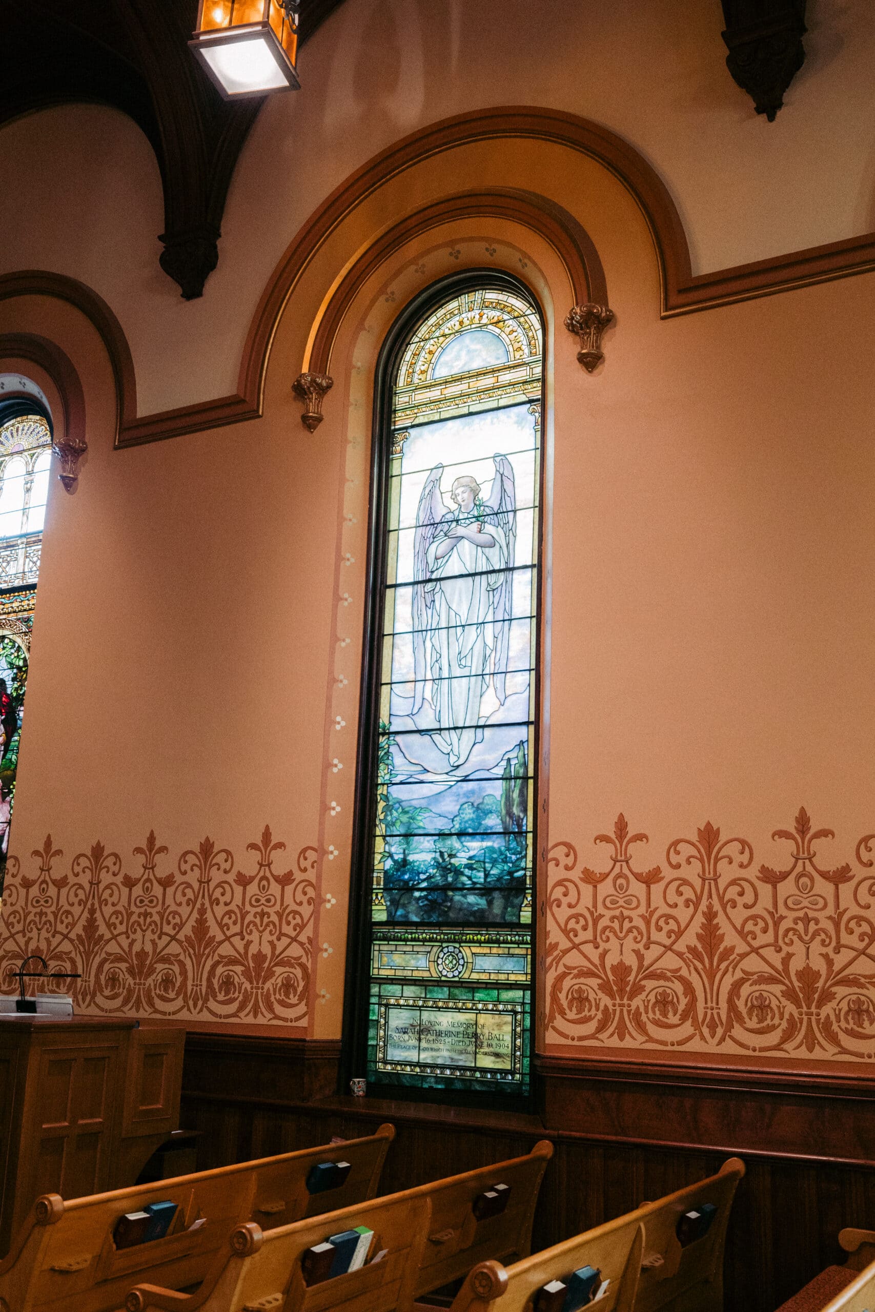 A tall stained glass window in a church wall depicts an angel with wings and a halo. The window is framed by ornate molding, and wooden pews are visible below it. The surrounding wall features decorative patterns. - McNatt Contracting