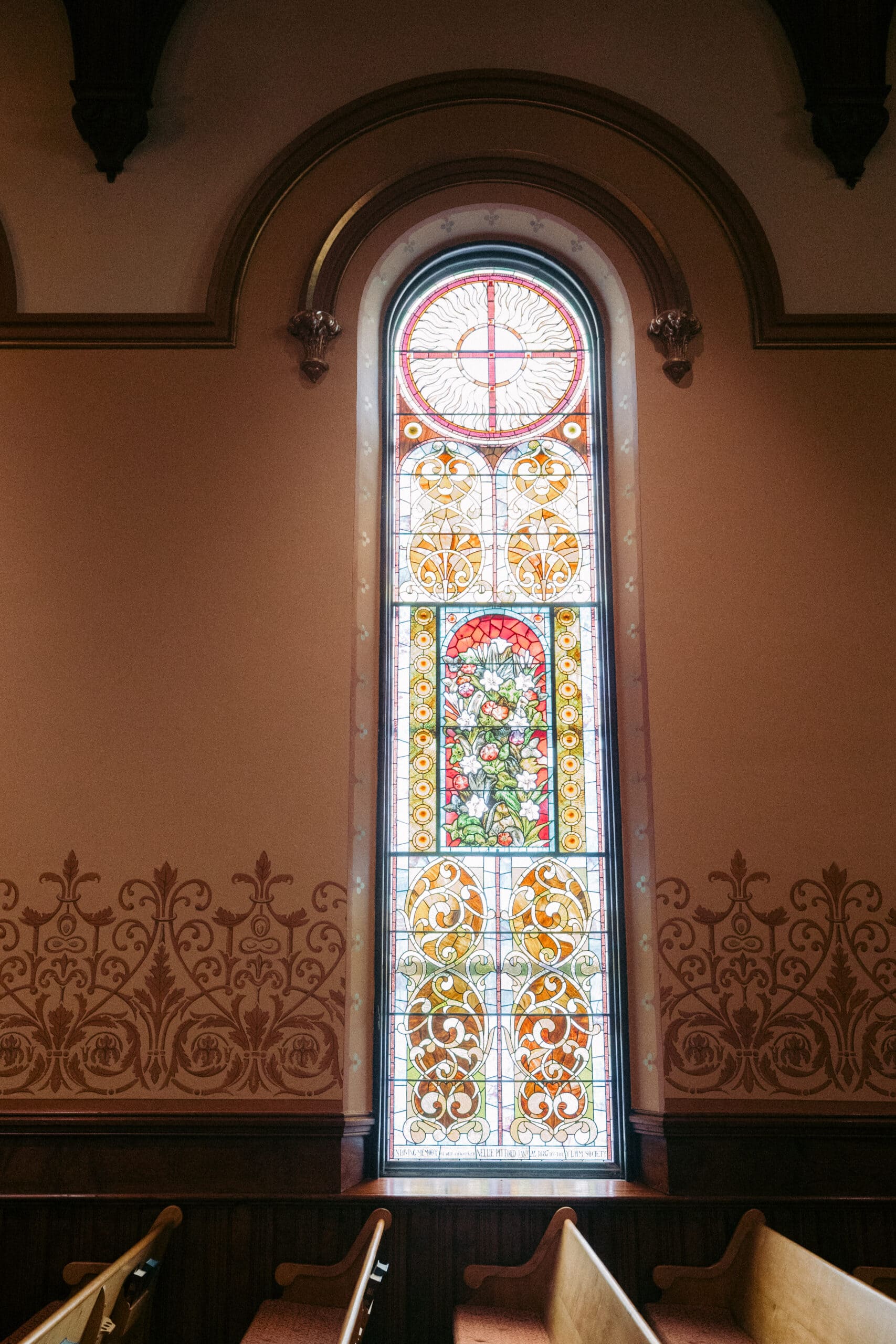 A tall arched stained glass window with floral and geometric designs is set in a beige wall with ornate trim and decorative patterns. Wooden pews are partially visible at the bottom. - McNatt Contracting