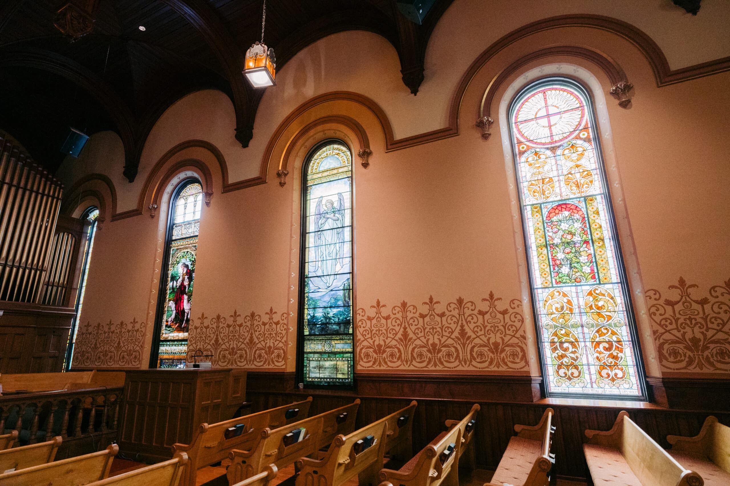 Interior of a church featuring wooden pews and three tall stained glass windows depicting various intricate designs and figures, with ornate wall detailing beneath each window. - McNatt Contracting