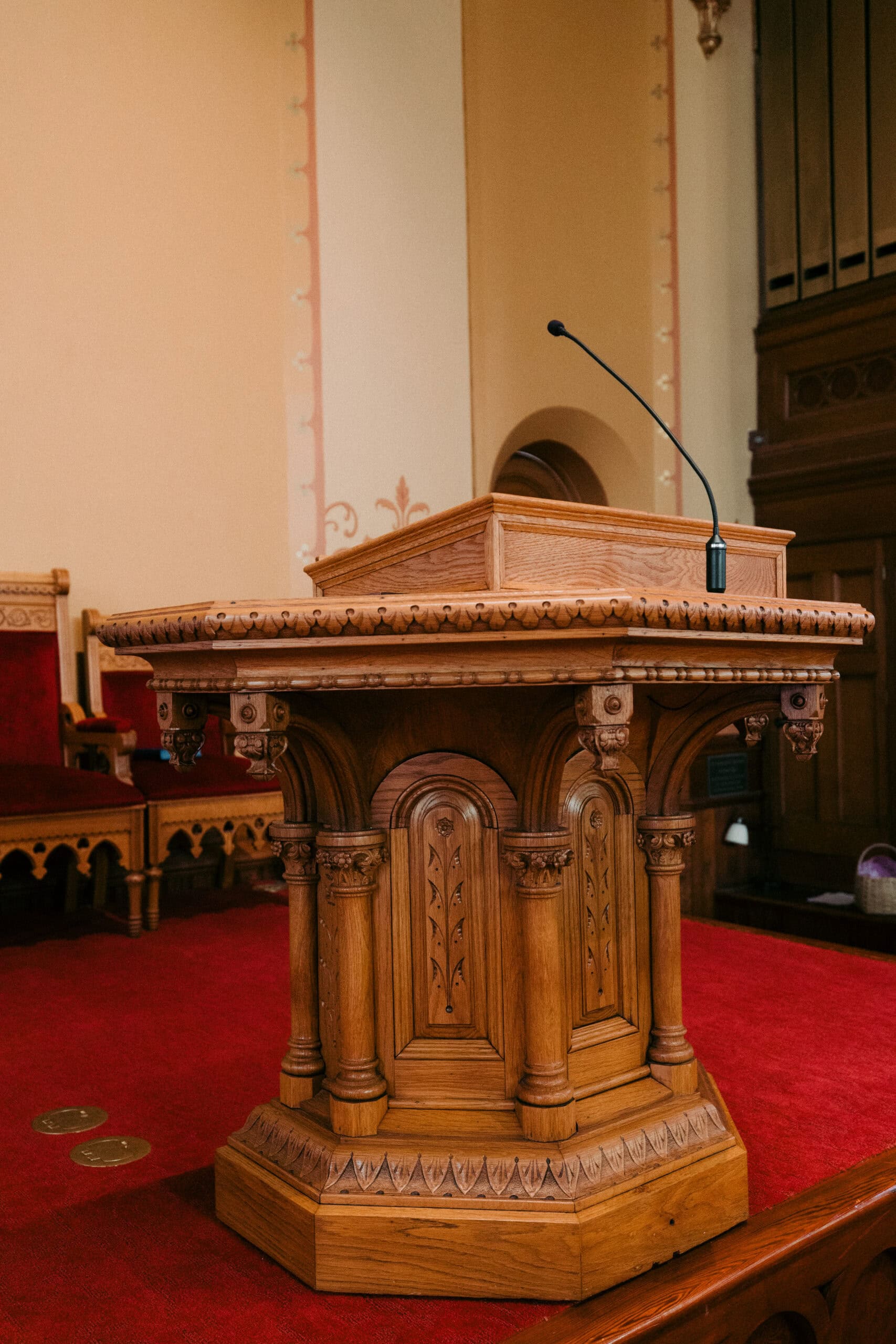A wooden podium with ornate carvings stands on a red carpeted platform. A microphone is mounted on top, and wooden chairs and paneling are visible in the background. - McNatt Contracting