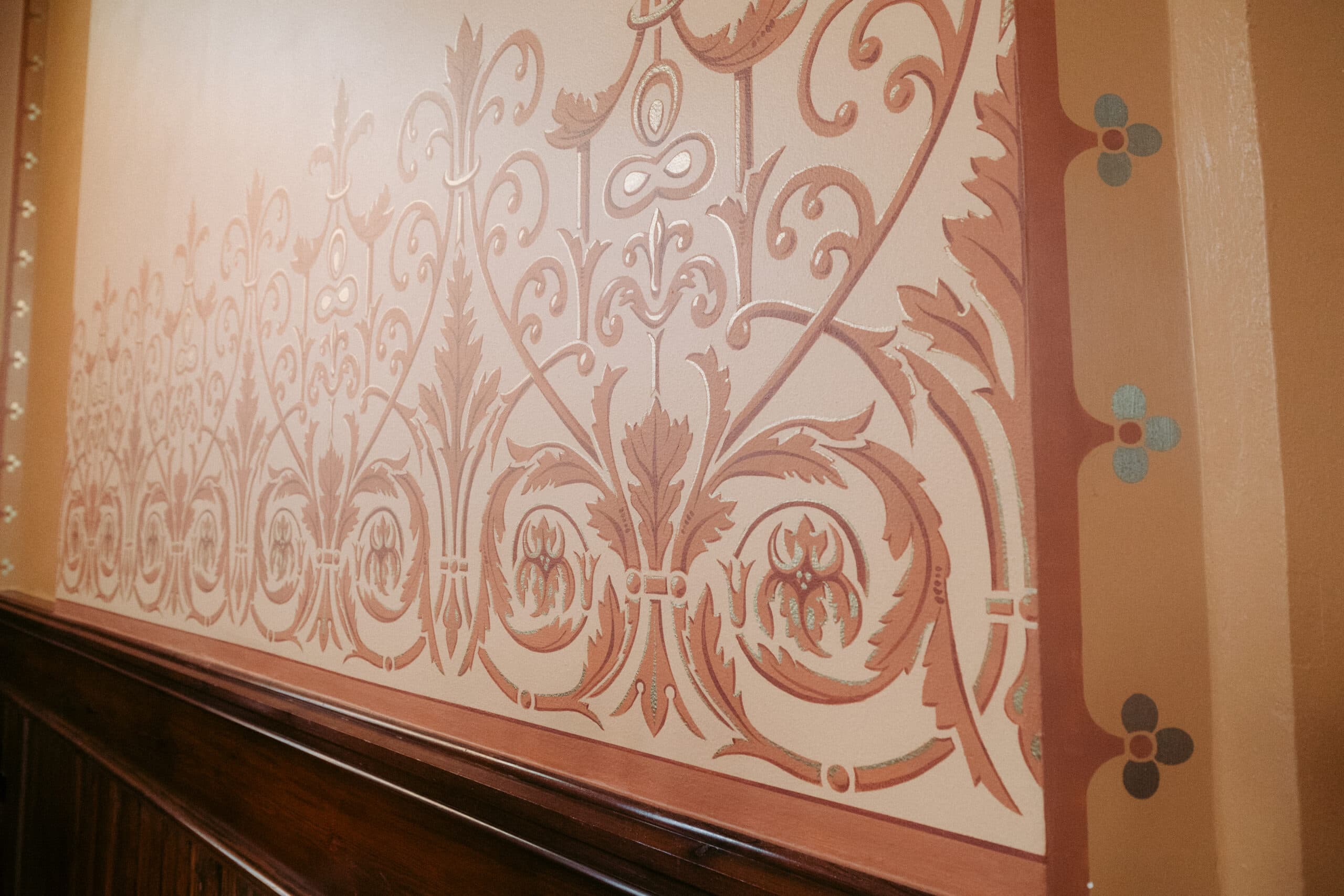 A wall with ornate brown and tan stenciled patterns, featuring swirling floral and leaf designs, bordered by a wooden trim at the bottom. The right edge shows a vertical band with small blue and red motifs. - McNatt Contracting