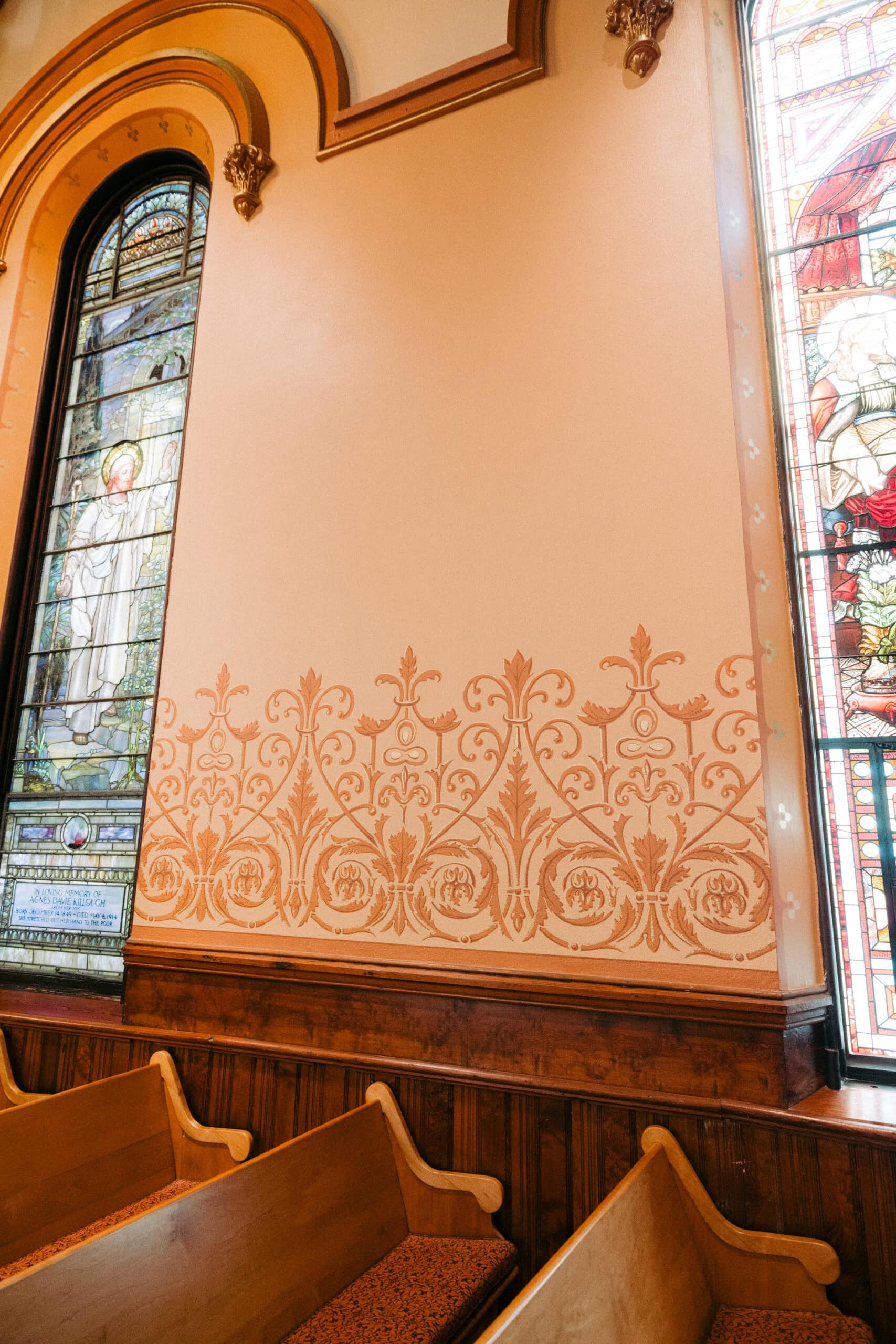 Wooden church pews sit in front of a peach-colored wall with ornate painted designs. The wall is flanked by tall stained glass windows depicting religious figures and scenes. - McNatt Contracting