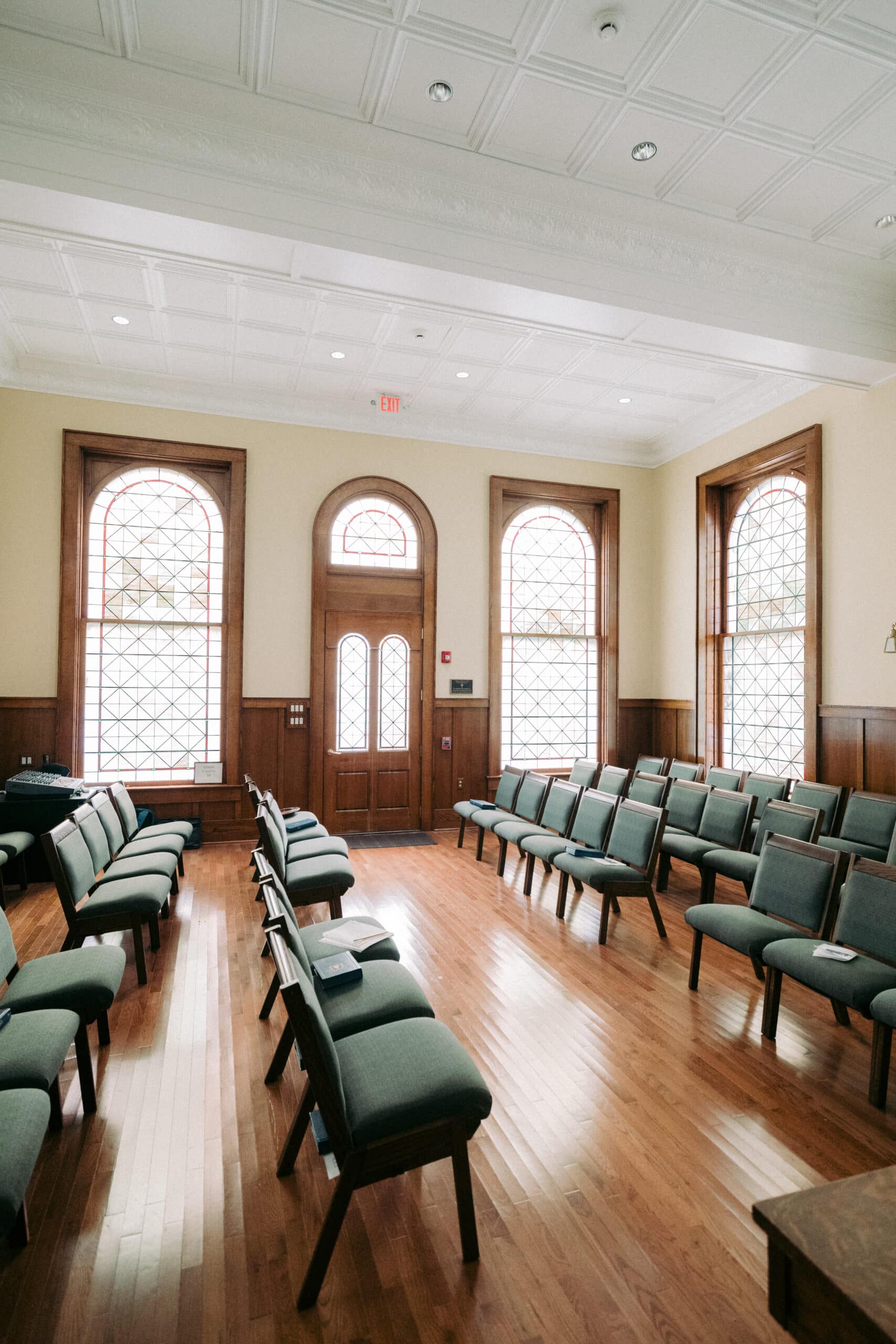 A room with rows of green cushioned chairs arranged on a hardwood floor, large arched windows with decorative grid patterns, and a wooden double door at the far end. - McNatt Contracting