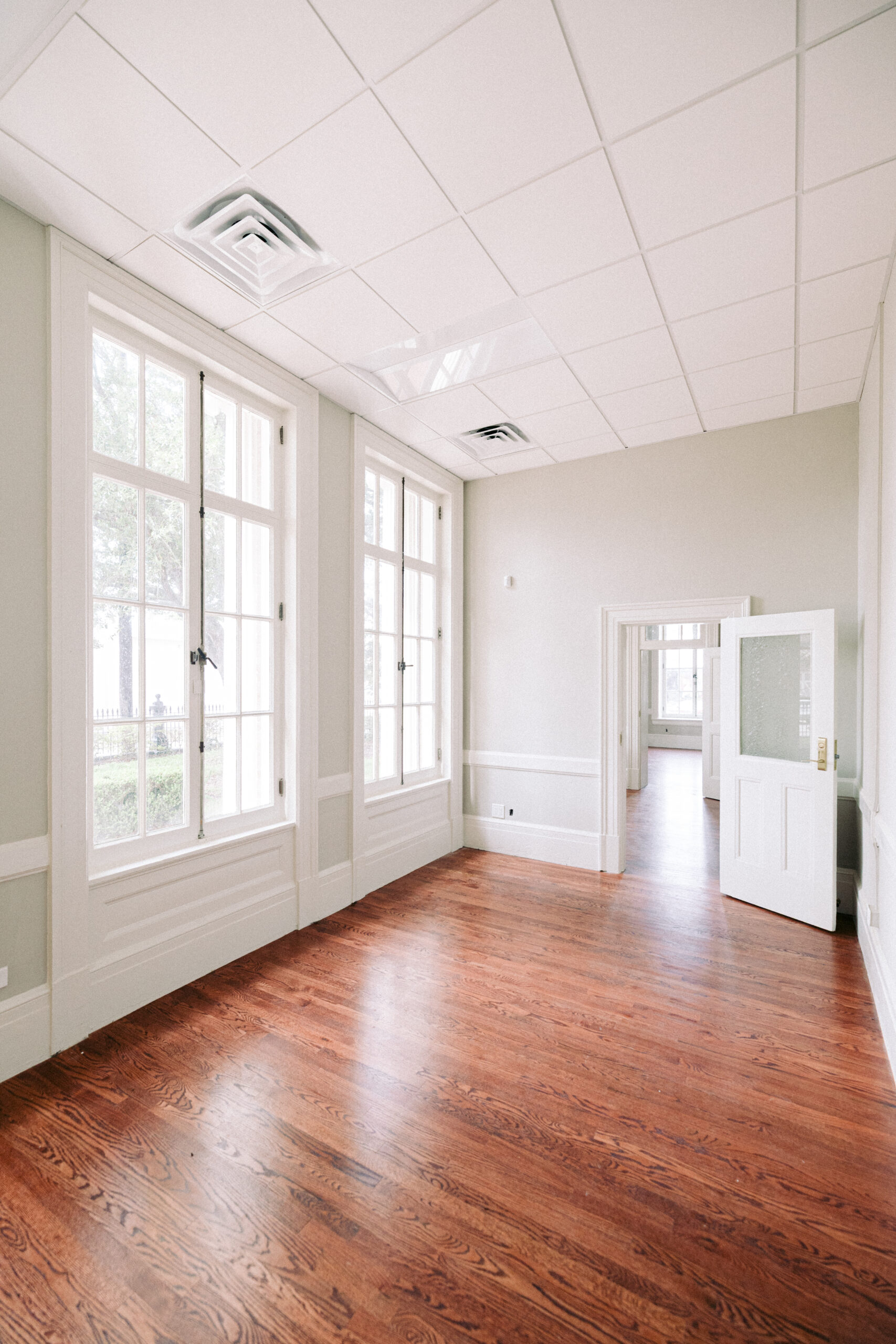 An empty room with light green walls, large white-trimmed windows, a white ceiling with panels, and polished wooden floors. An open door leads to another similar room in the background. - McNatt Contracting