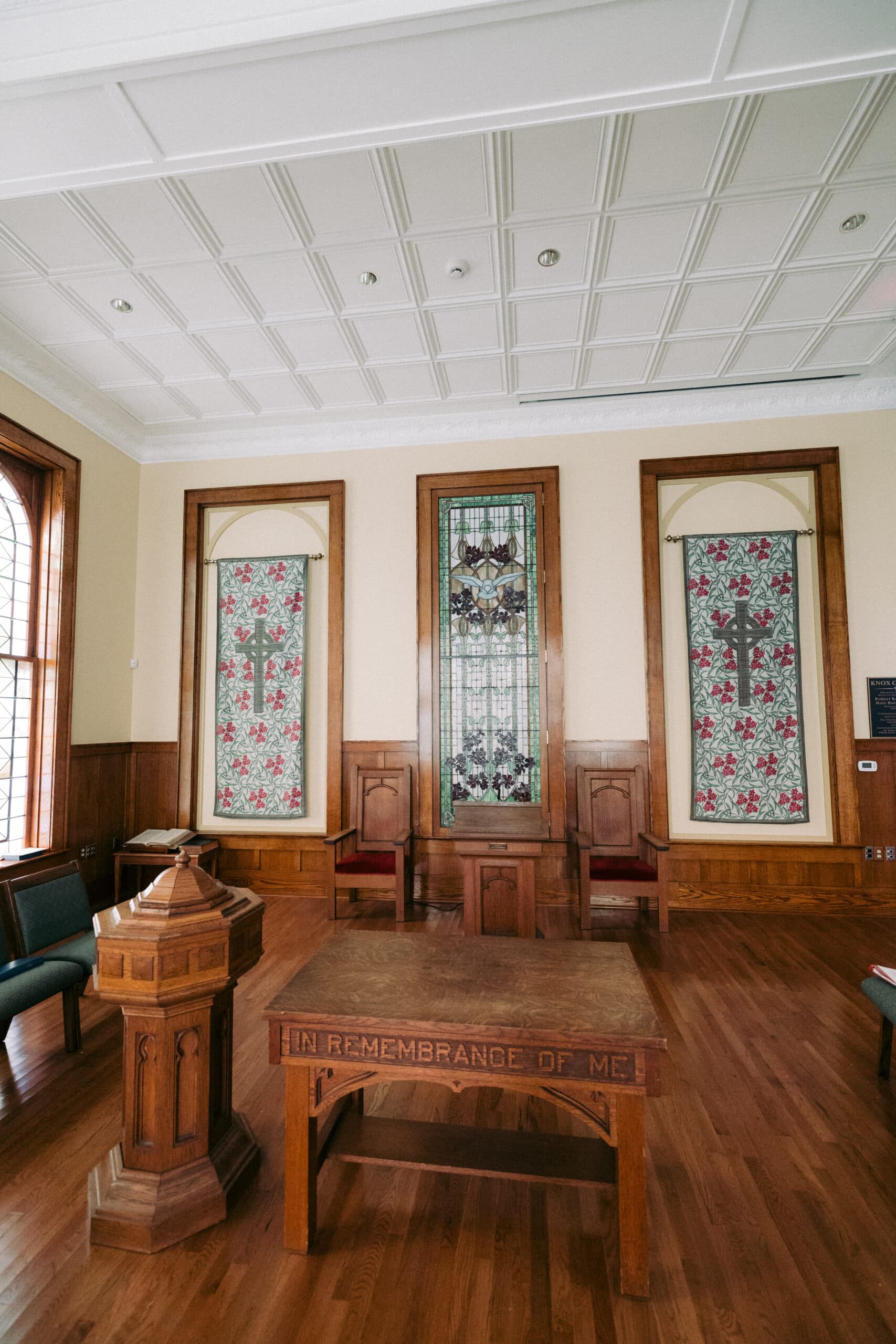 A wooden table with "IN REMEMBRANCE OF ME" carved on the side stands in a room with wood flooring, stained glass windows, and paneled walls. A wooden pulpit and chairs are also present. - McNatt Contracting