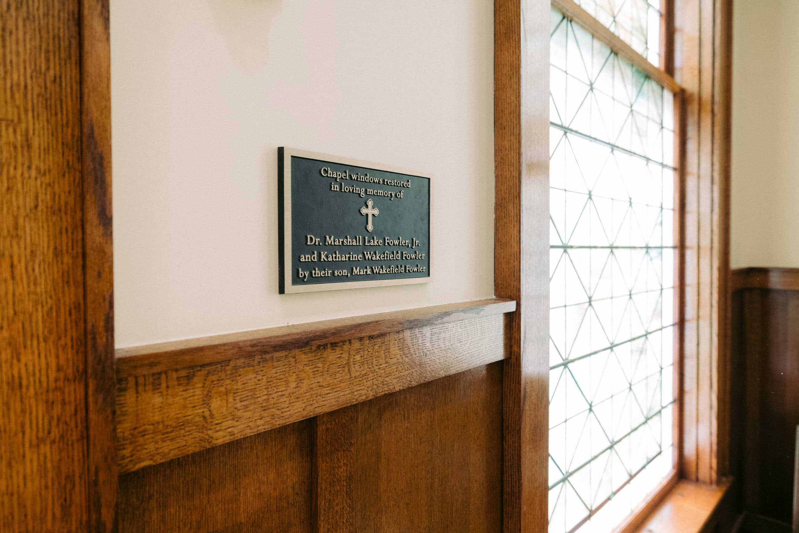 A metal plaque on a wooden wall honors Dr. Marshall Lake Rowley Jr. and Katherine Whitefield Rowley. A window with decorative glass panels is on the right, letting in natural light. - McNatt Contracting