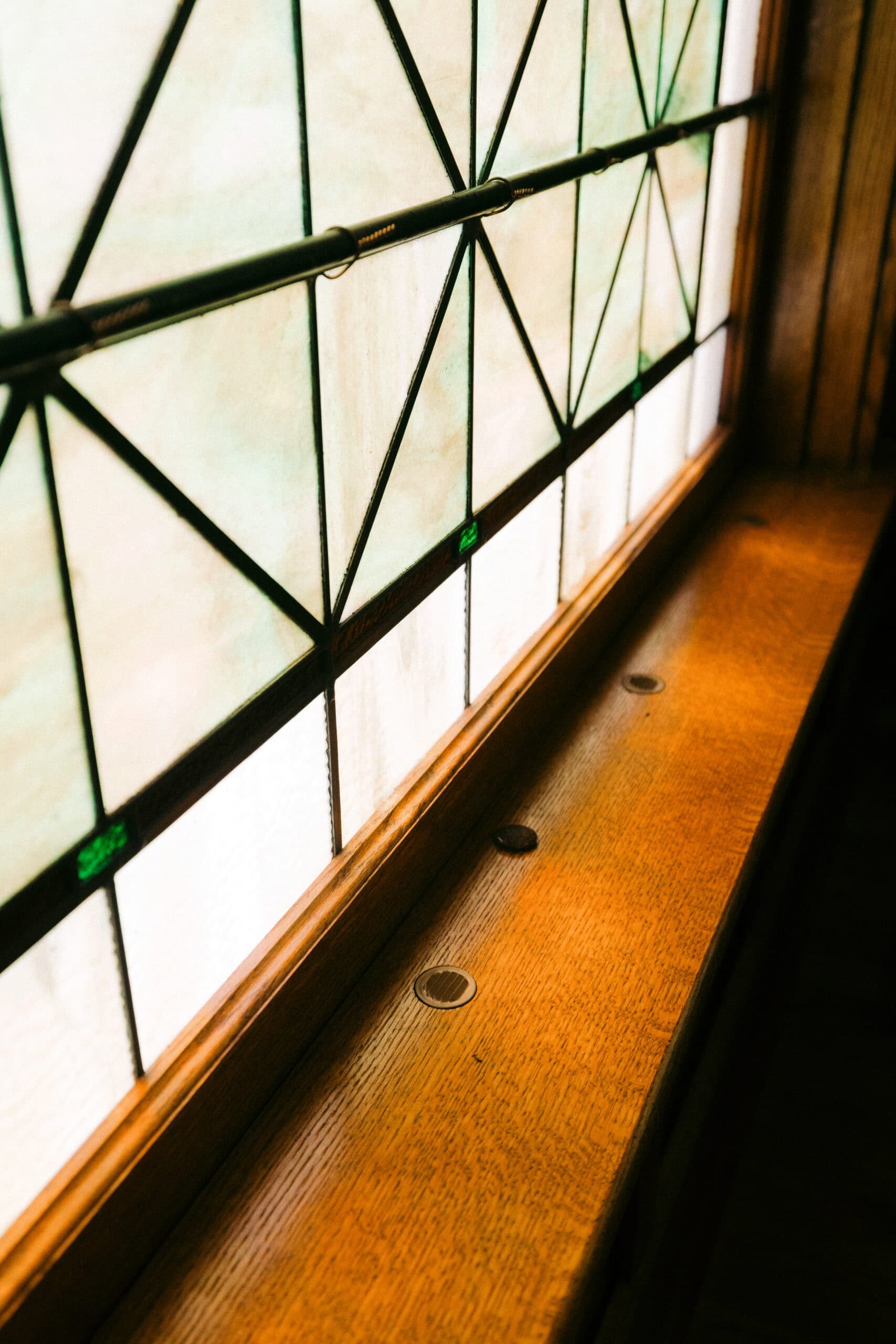 A close-up view of a wooden windowsill beneath a decorative stained glass window with geometric patterns and green accents. Sunlight filters through, illuminating the wood surface. - McNatt Contracting
