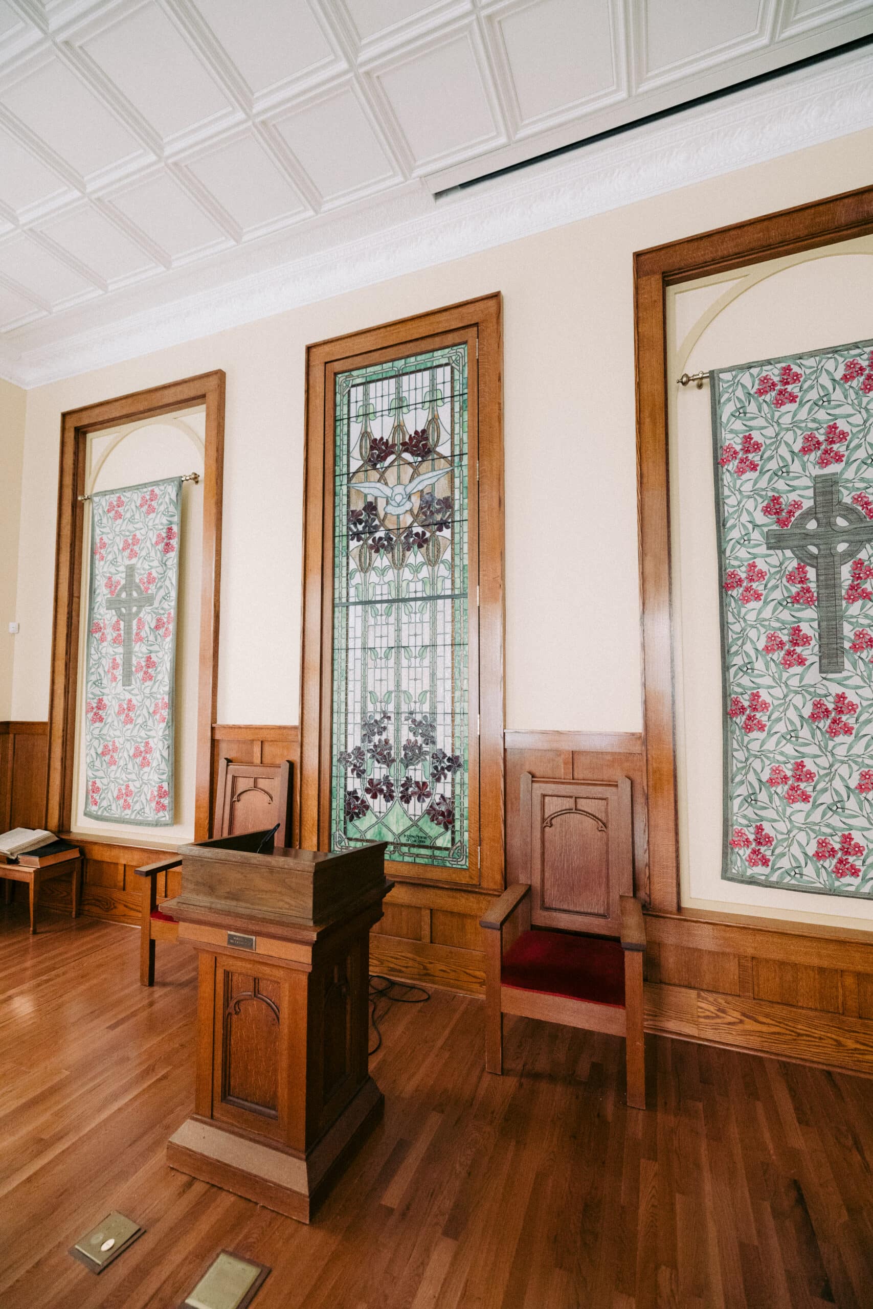 A wooden podium stands in front of three high-backed chairs on a hardwood floor. Behind them are tall stained glass and floral-patterned panels framed in wood, set against cream-colored walls. - McNatt Contracting