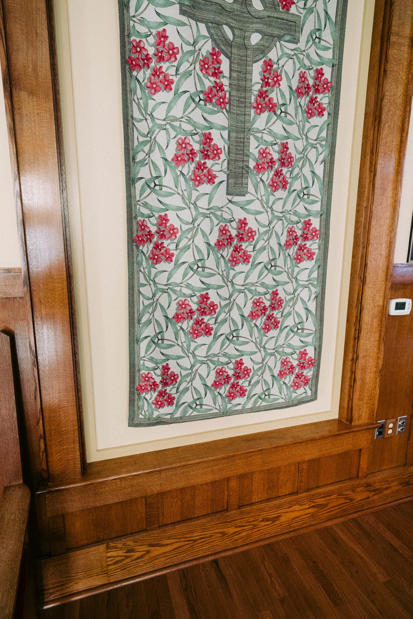 A rectangular textile wall hanging with a pattern of green vines and clusters of red flowers is framed by a wooden border and mounted on a cream-colored wall above a hardwood floor. - McNatt Contracting