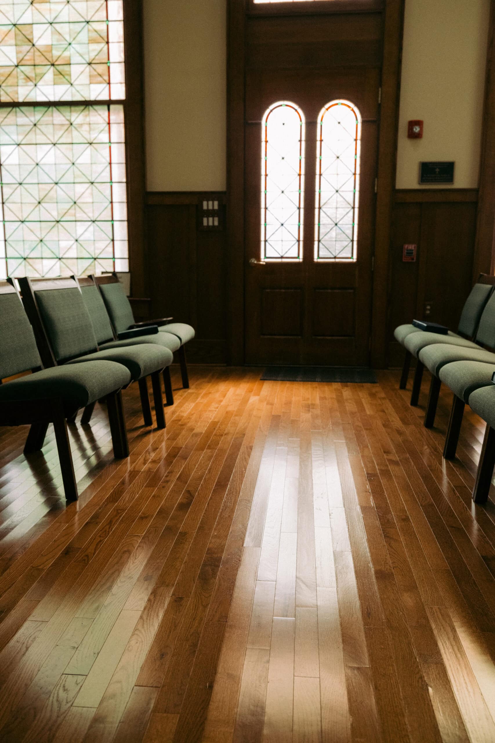 Rows of empty padded chairs line both sides of a polished wooden floor in a room with large windows and a double wooden door with arched glass panels. Light streams in, brightening the space. - McNatt Contracting
